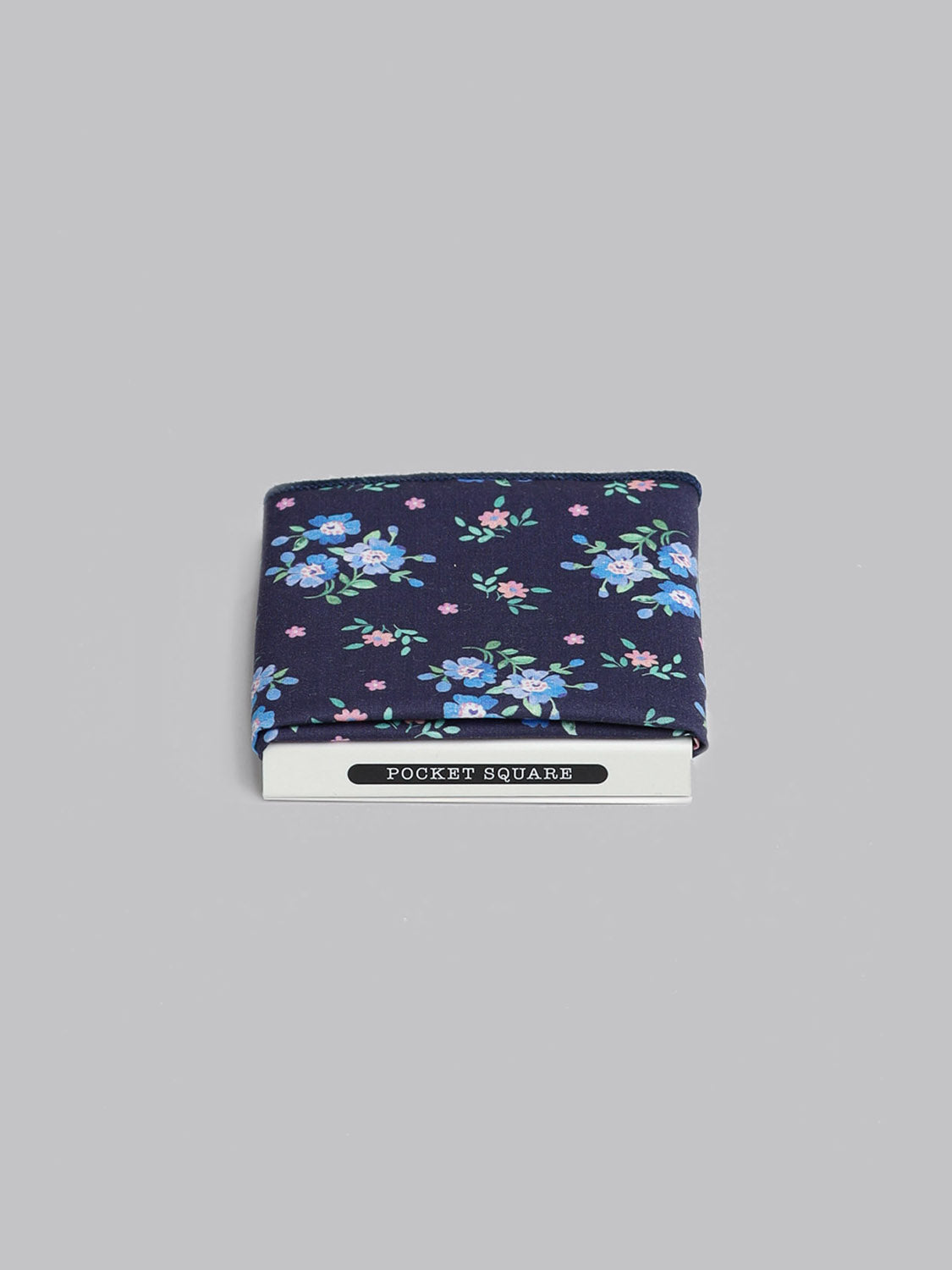 A Liberty™ Magdalena Pocket Square by ID Menswear, featuring a navy blue base with blue and pink floral print, is neatly folded in POCKET SQUARE packaging against a light gray background.