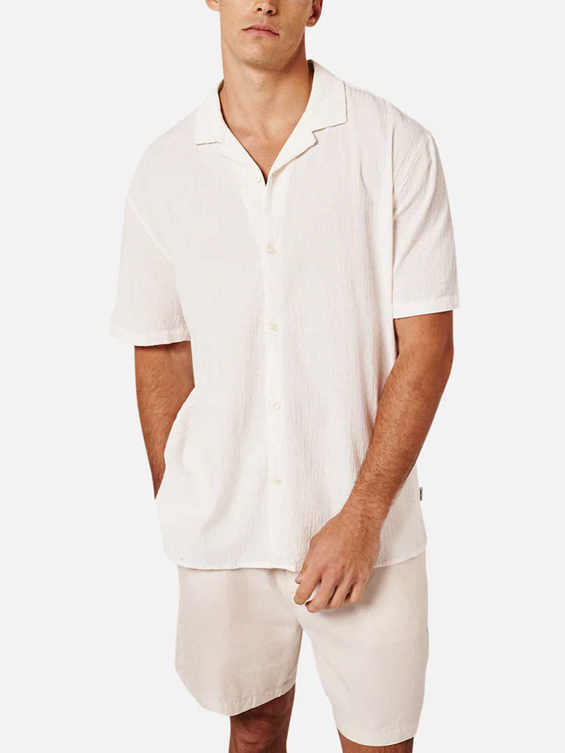 A man models The Magna Short Sleeve Shirt by Industrie—a breathable, off-white cotton top—paired with matching shorts. He stands with one hand in his pocket against a plain white background.