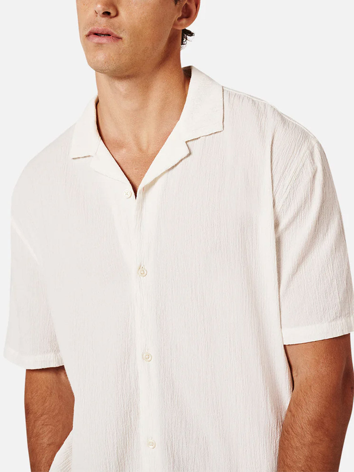 A man wearing Industrie’s Magna Short Sleeve Shirt—an oversized, white, breathable cotton shirt with a relaxed collar—stands against a plain light background. His face is partially out of frame.