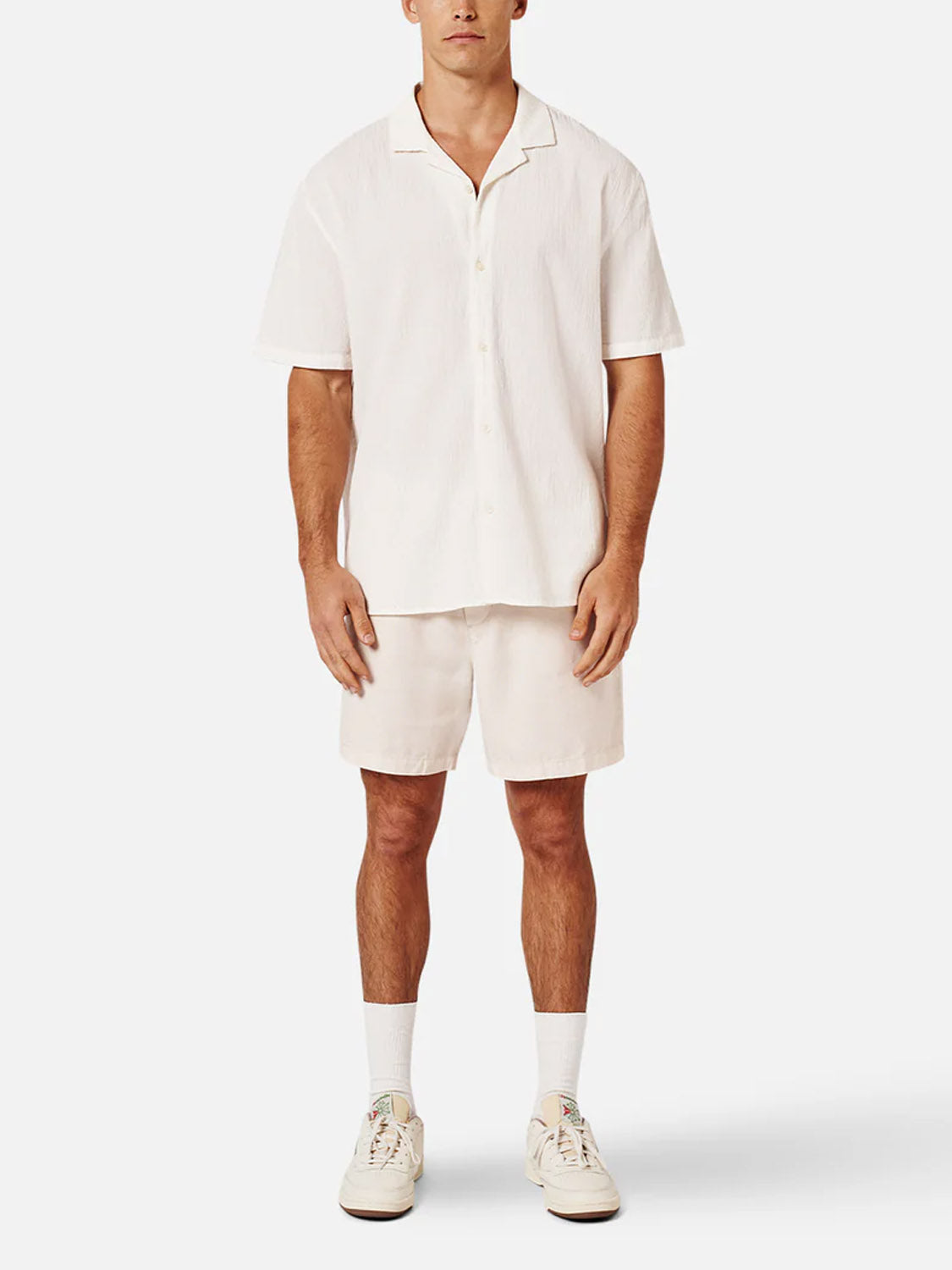 A man faces forward in Industrie’s The Magna Short Sleeve Shirt and matching off-white shorts, both in an oversized fit, paired with white sneakers and crew socks, set against a plain white background.