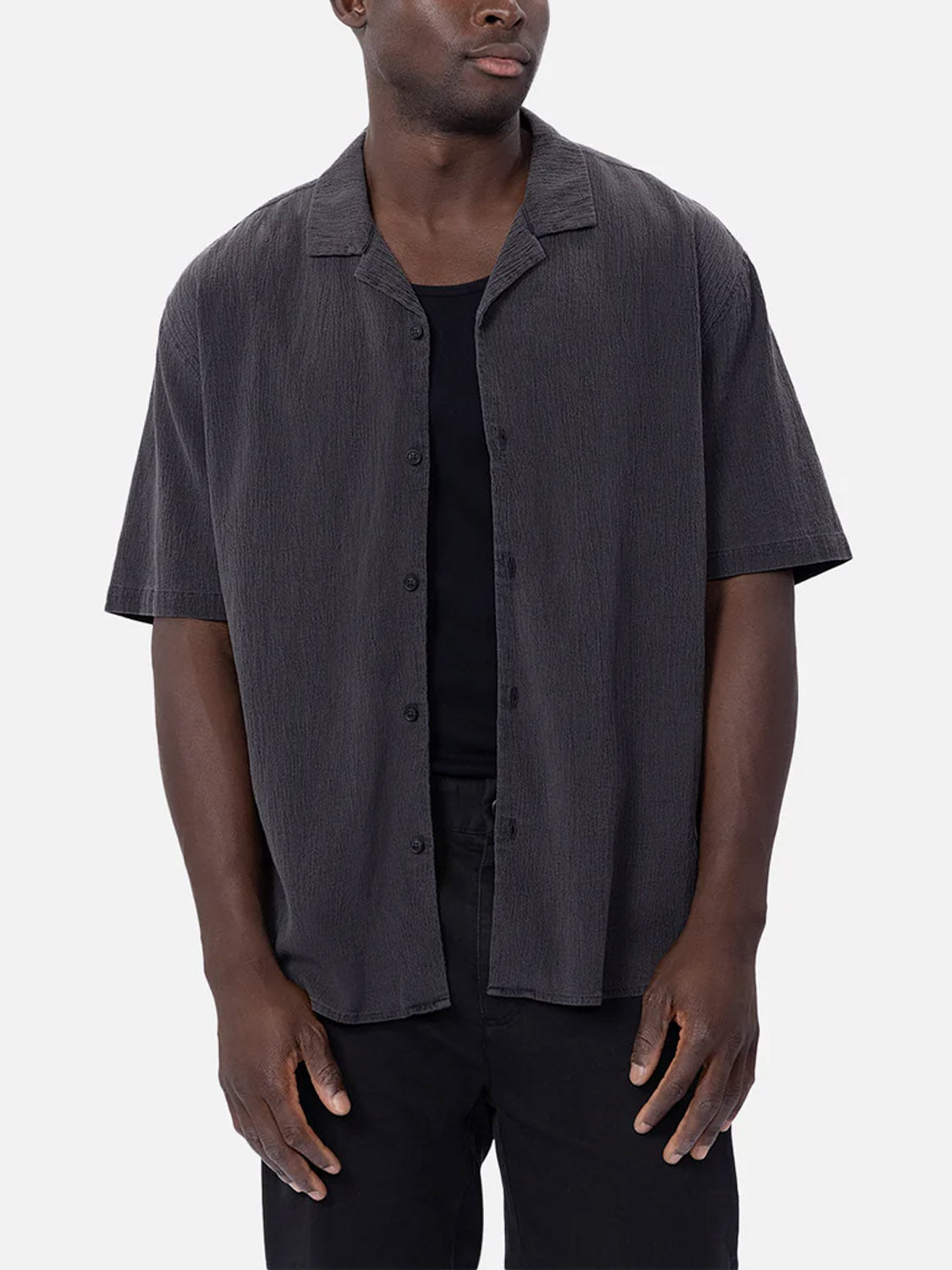 A man wears Industrie’s The Magna Short Sleeve Shirt, a loose-fitting, breathable cotton top, layered over a black T-shirt and black pants, standing against a plain light gray background.