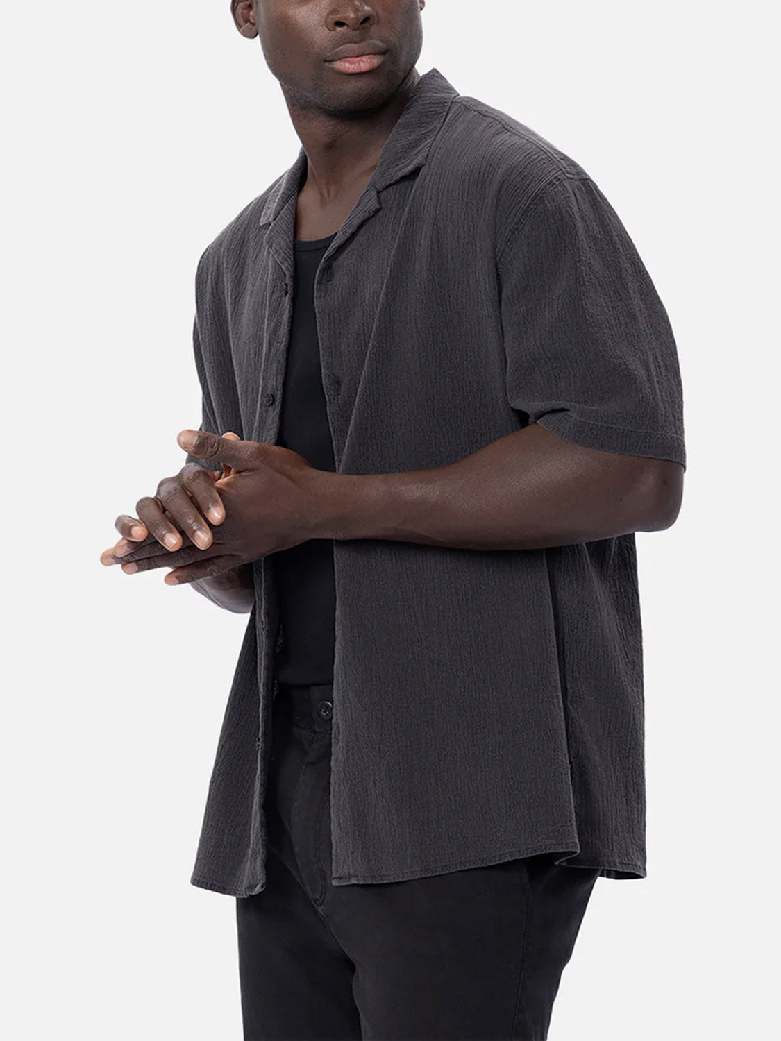 A man wears the Industrie Magna Short Sleeve Shirt in textured dark gray, styled oversized over a black t-shirt and black pants, standing against a plain light background with his hands clasped, looking to the side.