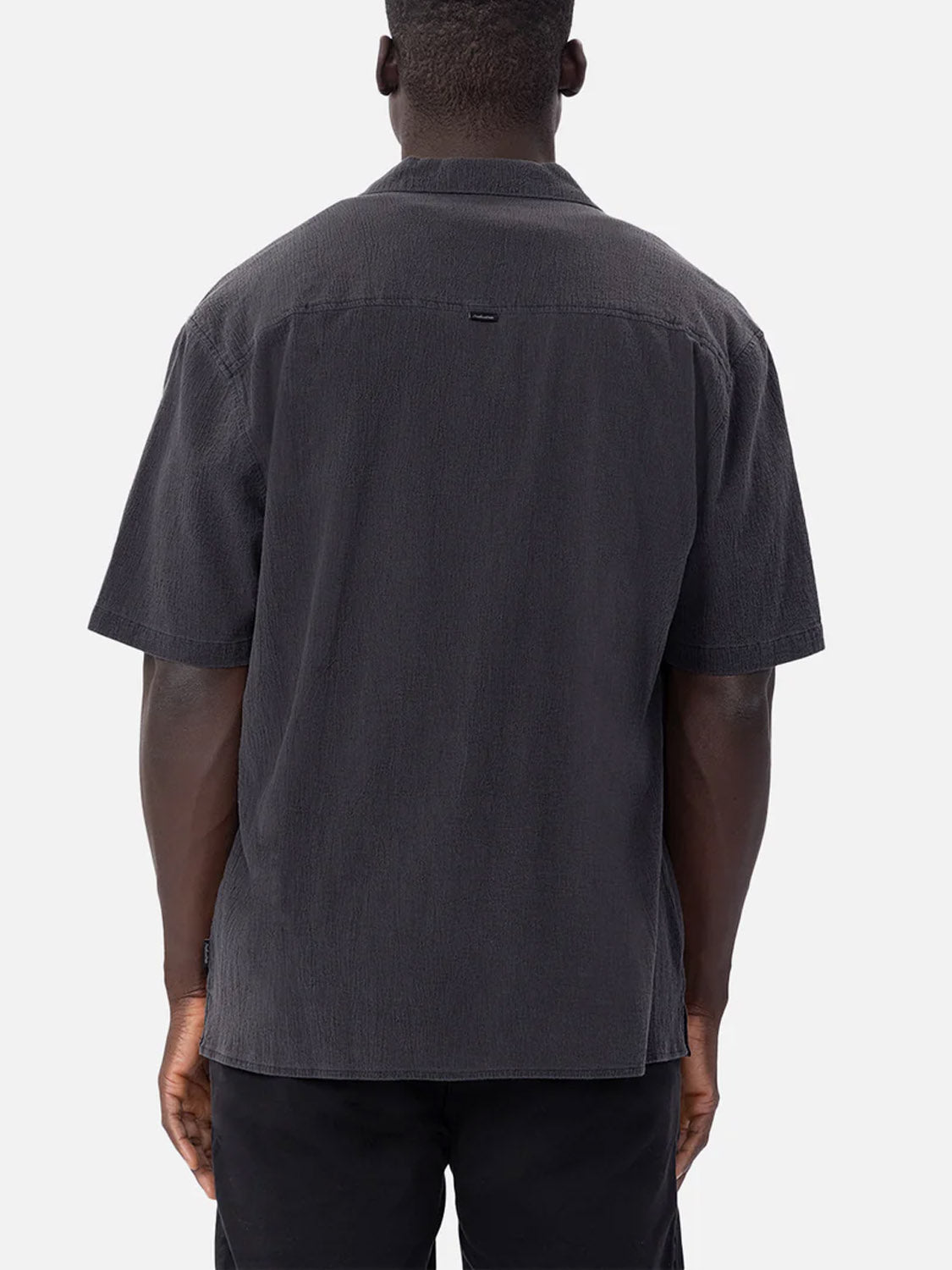 A person with short hair faces away from the camera, wearing Industrie’s The Magna Short Sleeve Shirt in black with an oversized fit and textured fabric, paired with black pants, set against a plain light background.