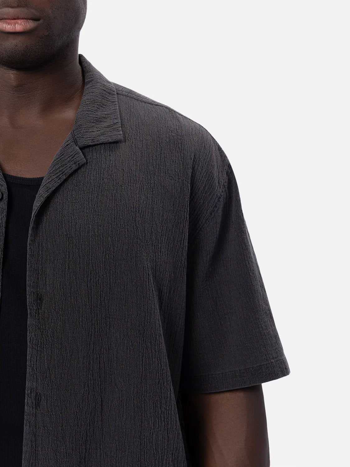 A person wears Industrie’s The Magna Short Sleeve Shirt in textured dark grey, oversized fit, layered over a black top and shown from shoulders to mid-torso against a plain light background.