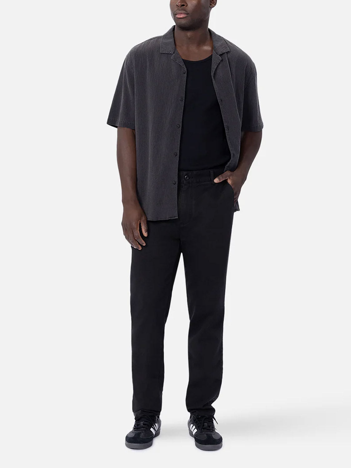 A man wears the Industrie Magna Short Sleeve Shirt—an oversized, breathable cotton piece—layered over a black tee with black jeans and black sneakers featuring white details. One hand in his pocket, he looks off to the side against a plain background.