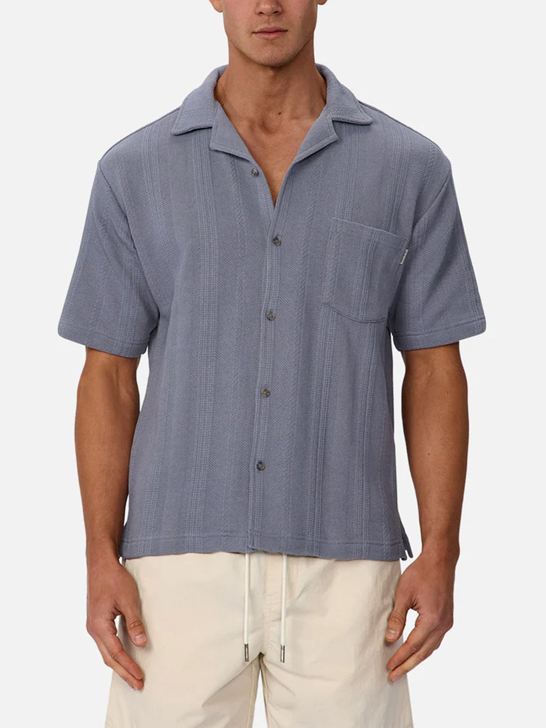 A man wears the Industrie Malaga Short Sleeve Shirt, a lightweight light blue camp collar shirt with a chest pocket, paired with white drawstring shorts, standing against a plain light background.
