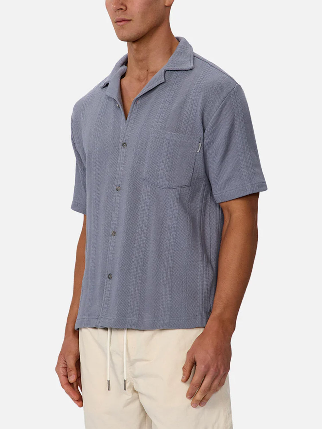A man wears the Industrie Malaga Short Sleeve Shirt, featuring light blue textured knit, short sleeves, and a chest pocket, paired with light drawstring pants—a standout lightweight piece against a plain background.