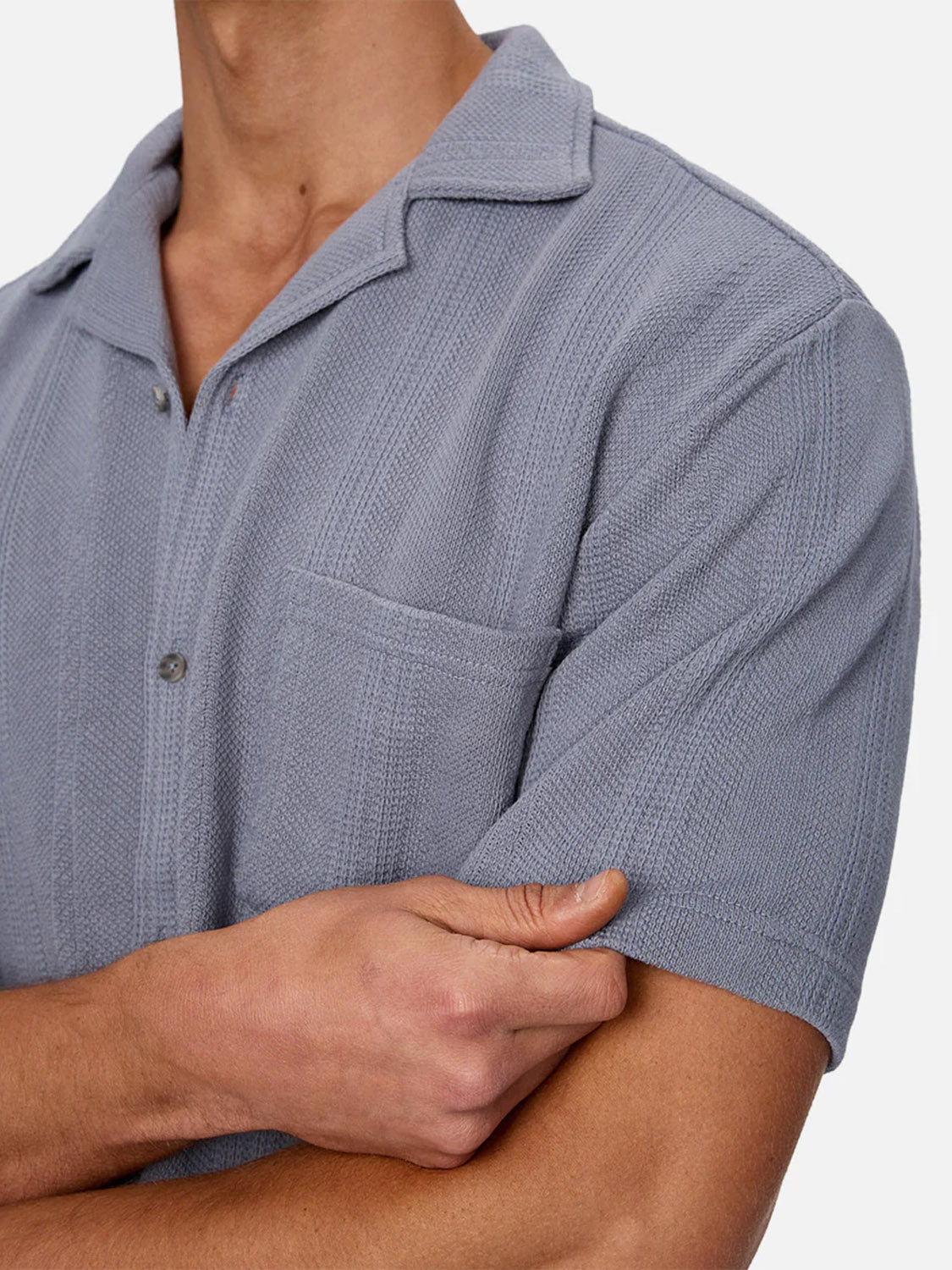 A person models the Industrie Malaga Short Sleeve Shirt—light grey, textured knit with a camp collar and chest pocket. With one arm crossed, their head partly out of frame, the focus is on this standout lightweight piece.