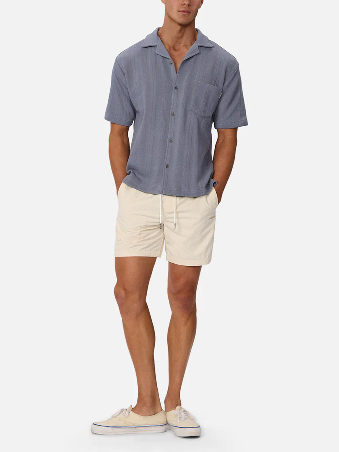 A man stands with hands in pockets, wearing Industrie’s Malaga Short Sleeve Shirt in blue, paired with light beige drawstring shorts and beige sneakers against a plain light background.