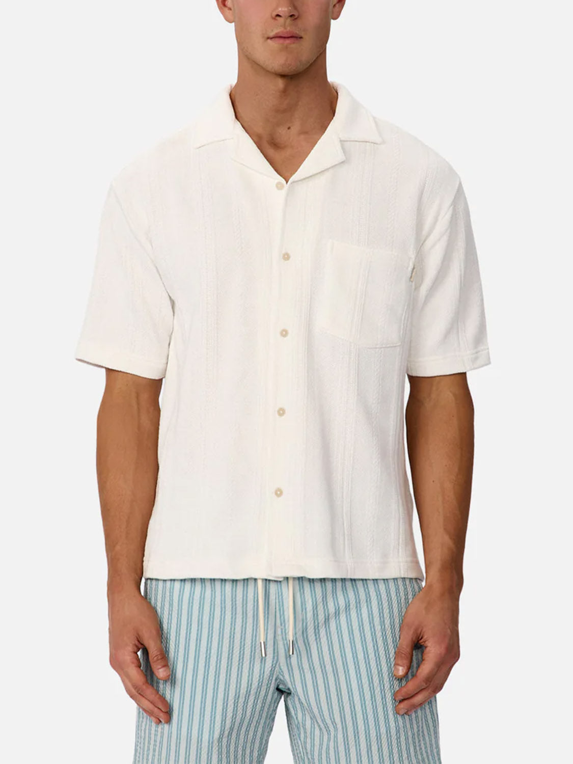 A man wears the Industrie Malaga Short Sleeve Shirt—a lightweight, white camp collar shirt with a chest pocket—paired with light blue striped drawstring shorts, standing against a plain white background.