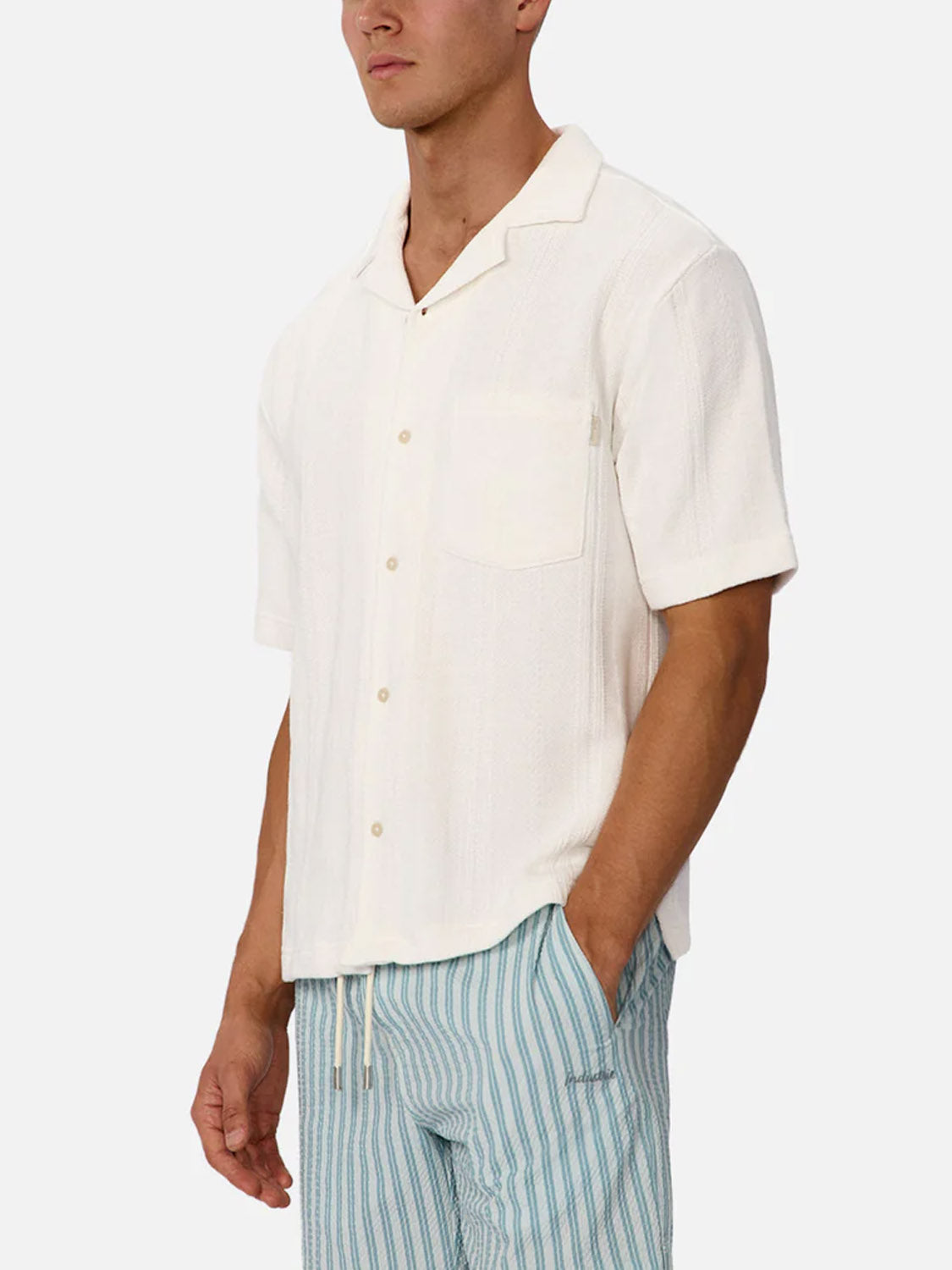 A man wears the Industrie Malaga Short Sleeve Shirt—an off-white camp collar shirt with a chest pocket—and light blue and white striped drawstring shorts, standing with one hand in his pocket against a plain background.