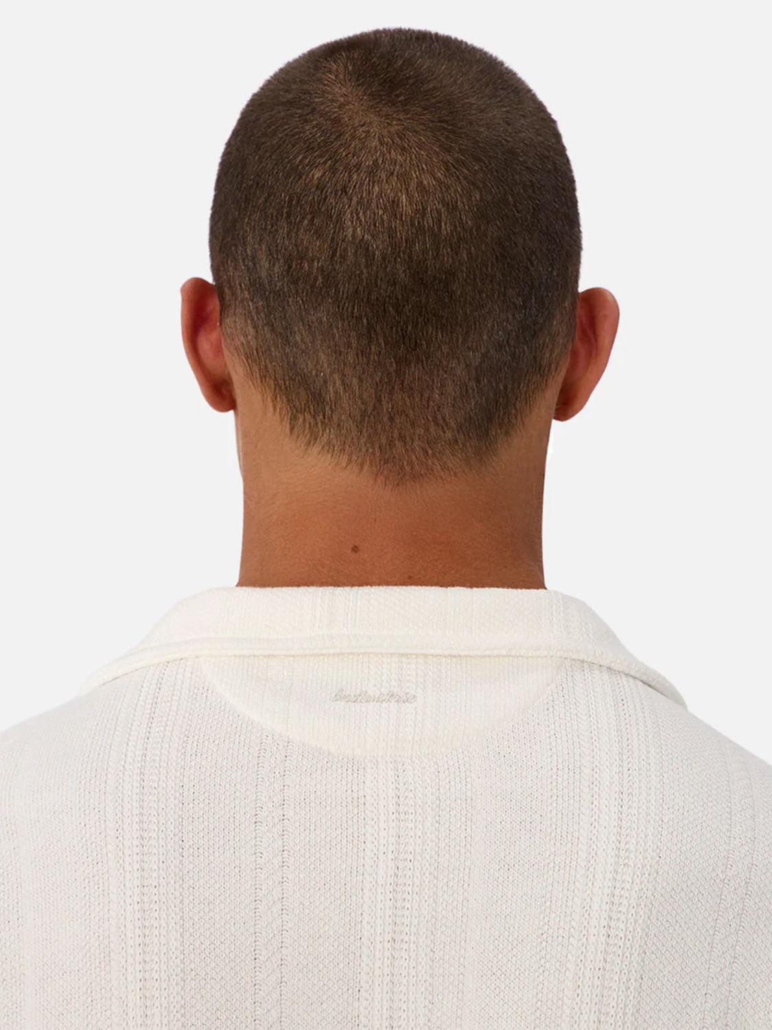 A person with short brown hair is seen from behind wearing the Industrie Malaga Short Sleeve Shirt, a white textured knit top with a collar and seam details, standing out against a plain, light background.