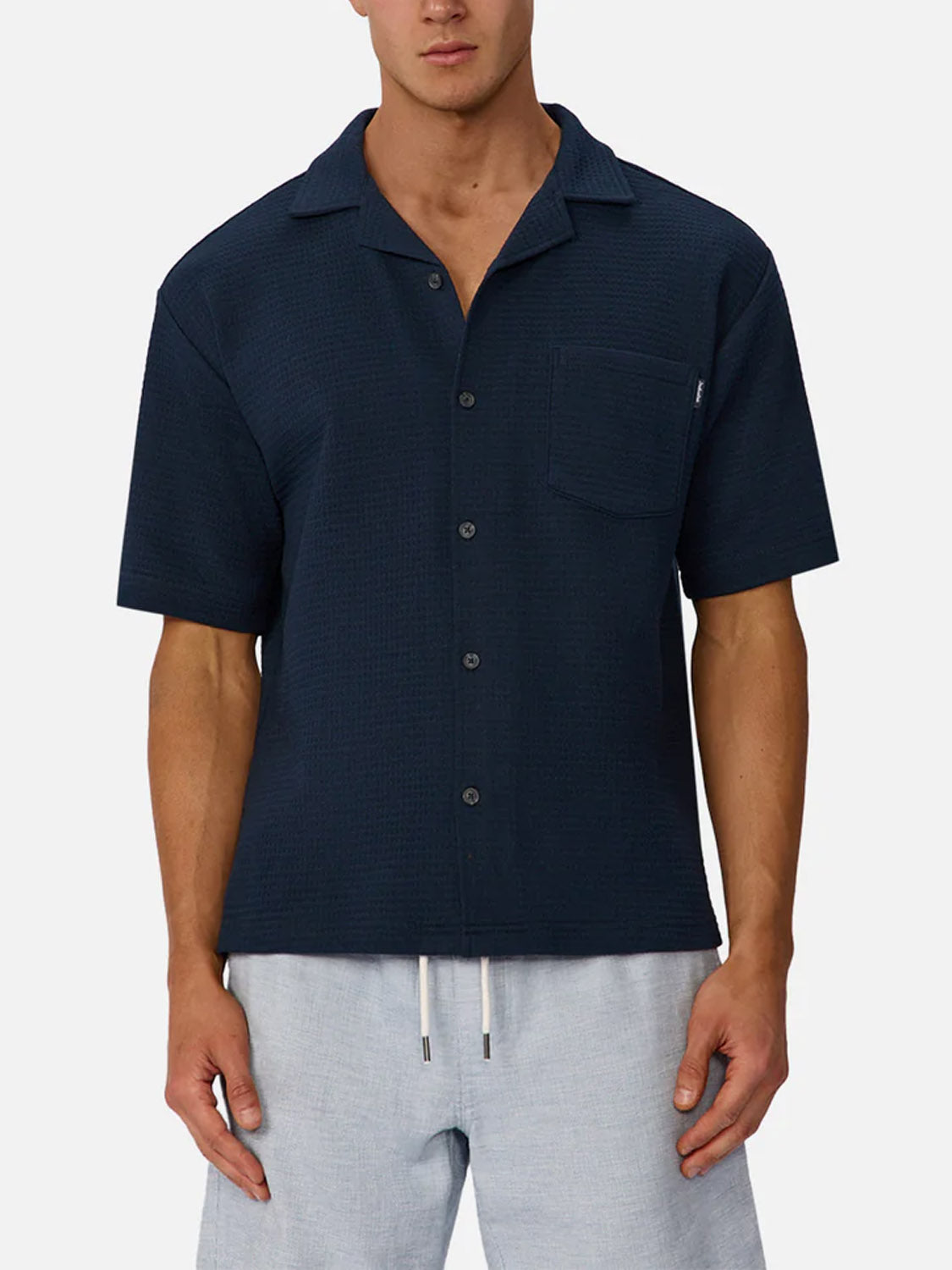 A man wears The Malibu Short Sleeve Shirt by Industrie in breathable cotton with a chest pocket, paired with light gray drawstring shorts, standing against a plain white background.