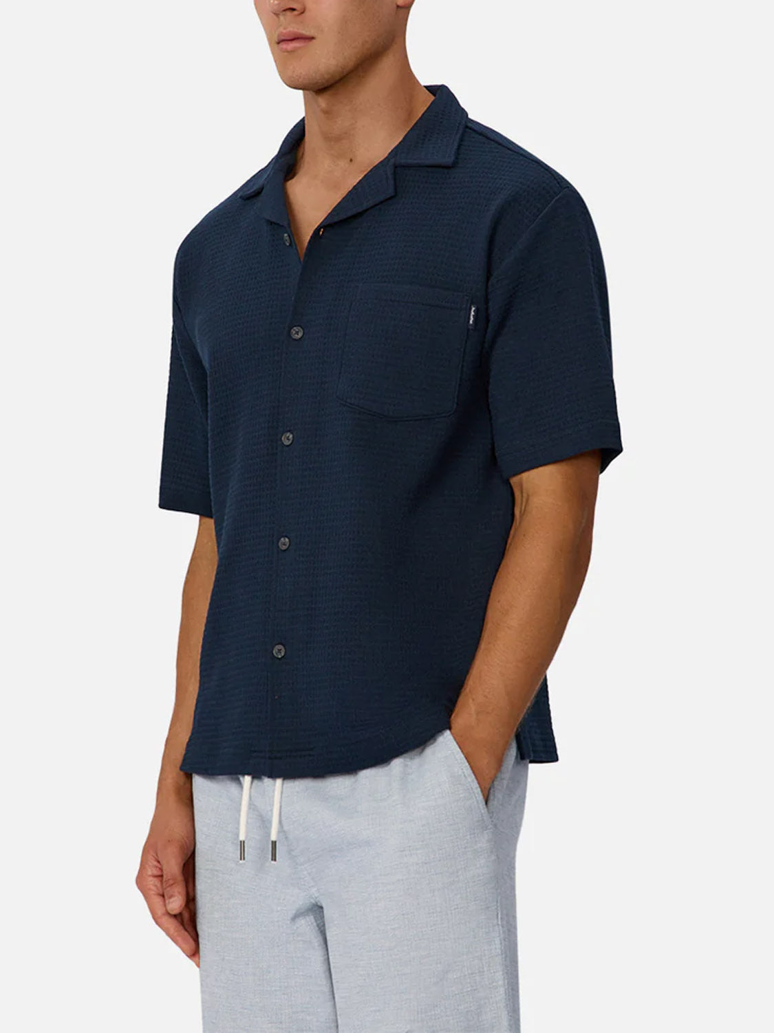A man wears the Industrie Malibu Short Sleeve Shirt, a breathable cotton top with a chest pocket, paired with light gray drawstring pants. He stands against a plain background with his face partially out of frame.