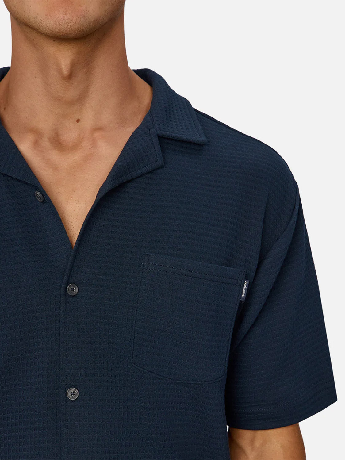 A person wears The Malibu Short Sleeve Shirt by Industrie—a dark navy, knitted button-up with a collar and chest pocket. The image is cropped at the nose, showing the upper torso against a plain background.