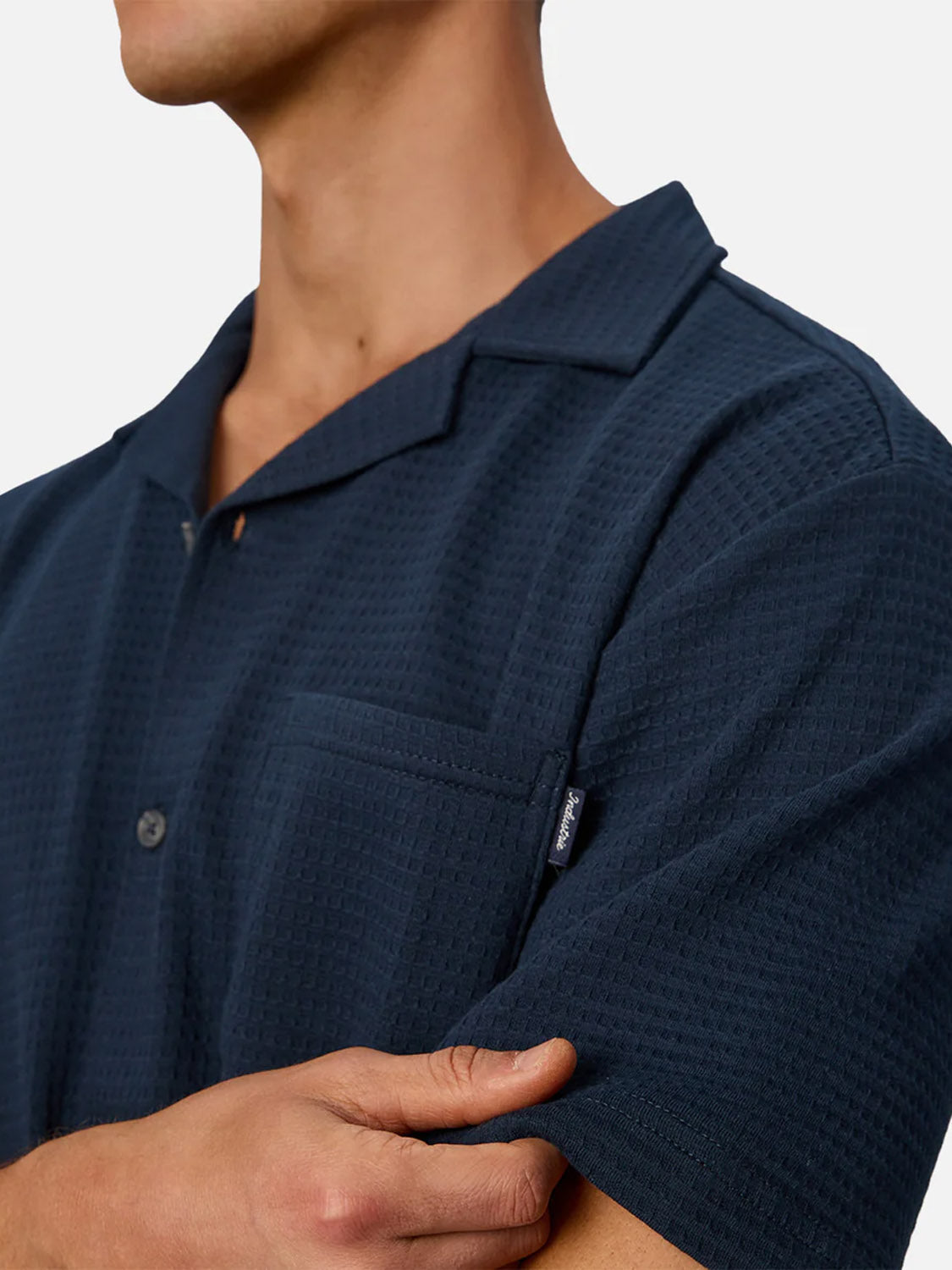 A person in The Malibu Short Sleeve Shirt by Industrie, featuring a dark blue knitted texture and chest pocket, stands with one arm crossed over their body; their face is partially out of frame.
