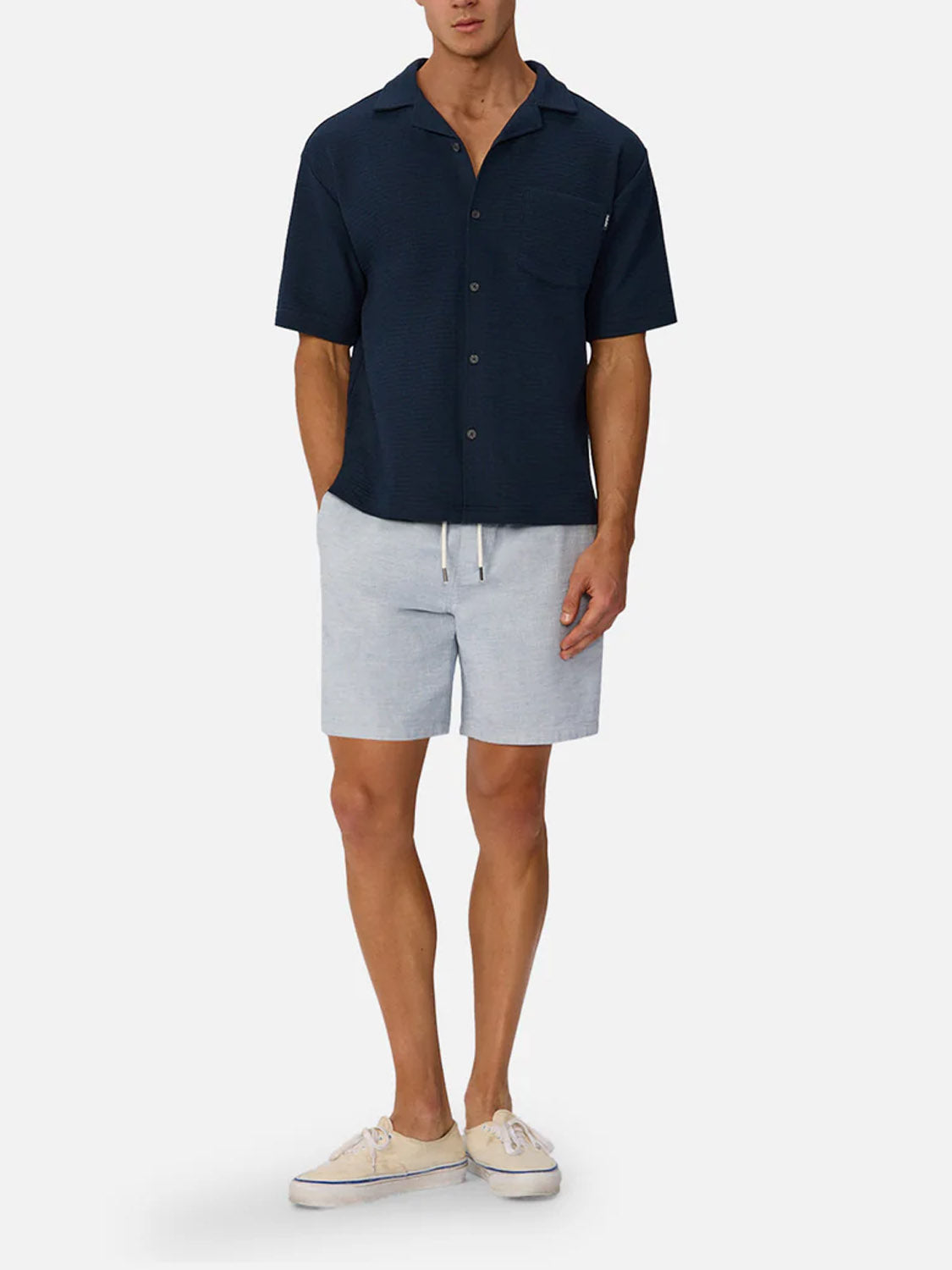 A man wears Industrie’s The Malibu Short Sleeve Shirt in navy, styled with light gray drawstring shorts and beige sneakers. Hands in pockets, he showcases the shirt’s breathable cotton knit against a plain white backdrop.