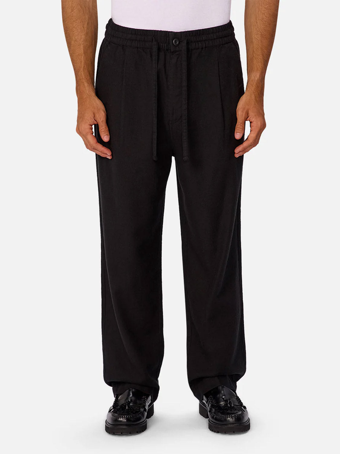 A person wears Industrie’s The Relaxed Mallorca Pant in black, featuring a relaxed fit, cotton fabric, and drawstring waist, paired with black shoes. Shown from the waist down against a plain light background.