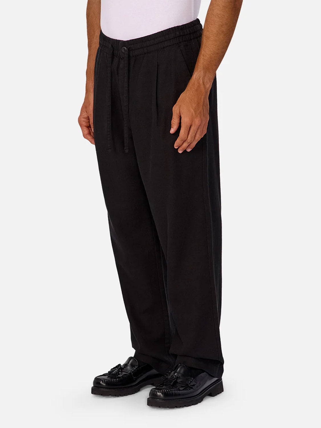 A person in a white shirt and Industrie’s The Mallorca Pant in black stands at an angle, hands by their sides. The breathable cotton pants are paired with black dress shoes against a plain, light background.