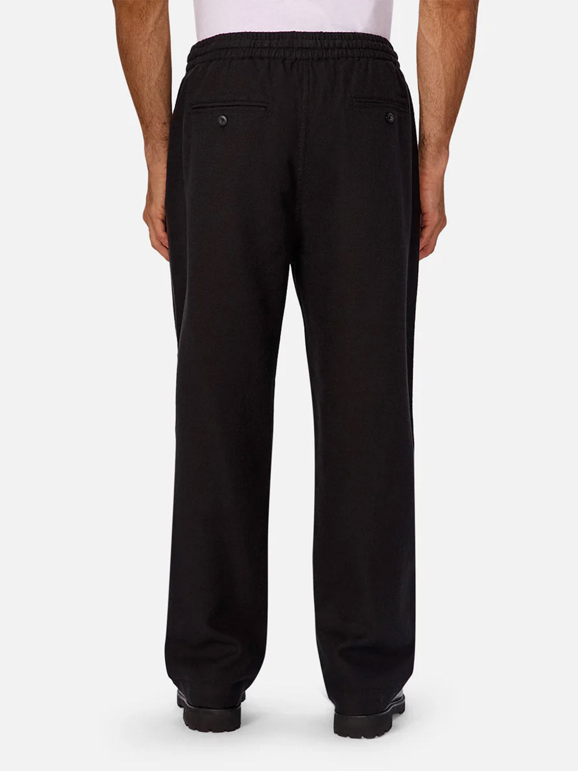 A person wearing Industrie’s The Mallorca Pant in black, featuring a relaxed fit, elastic waistband, and buttoned back pockets. Crafted from breathable cotton for all-day comfort, they are standing and facing away from the camera.