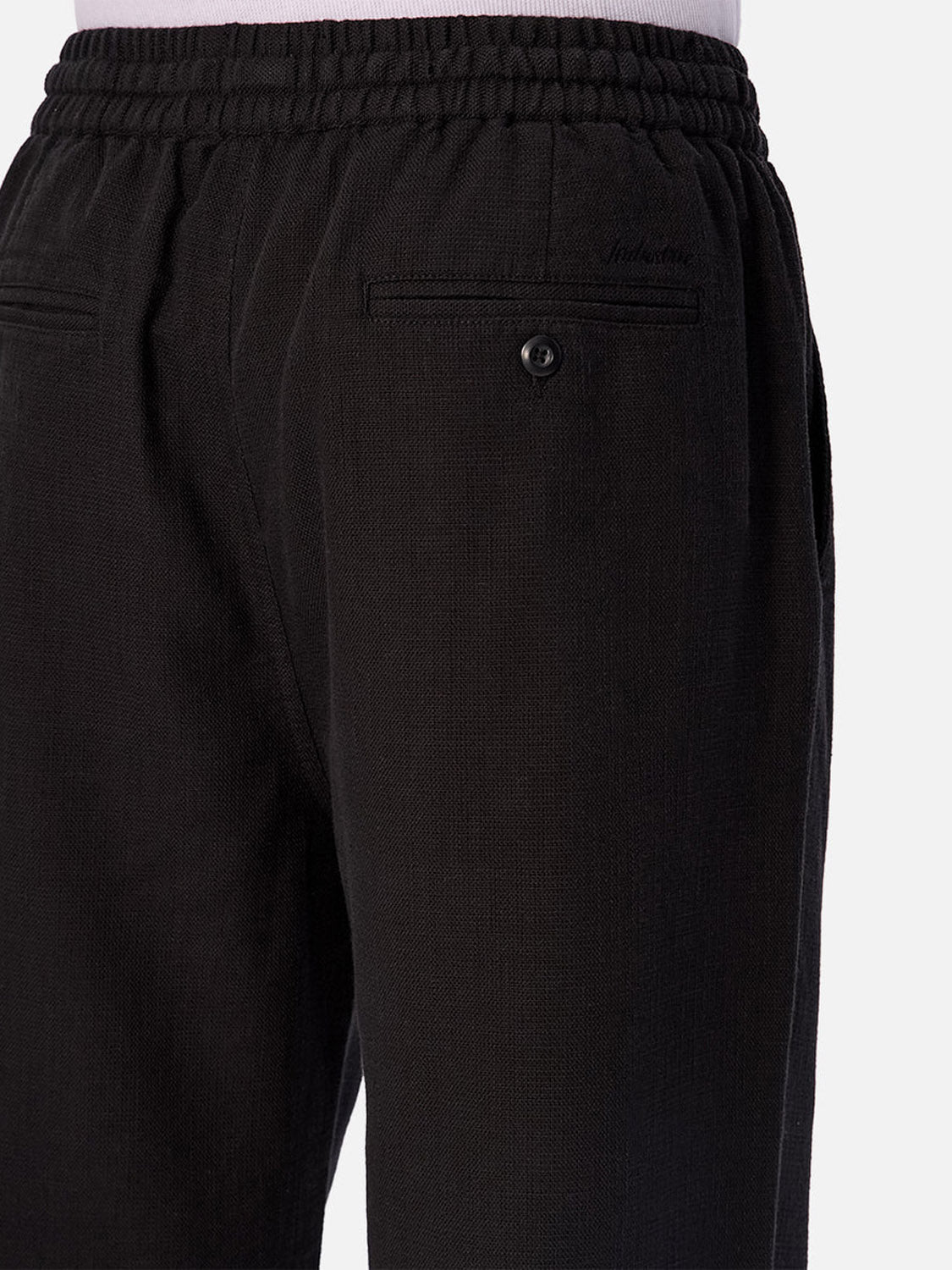 Close-up of a person wearing The Mallorca Pant by Industrie. Made from breathable black textured cotton, these pants have an elastic waistband and back and side pockets, including one with a button. A light-colored top is tucked in.