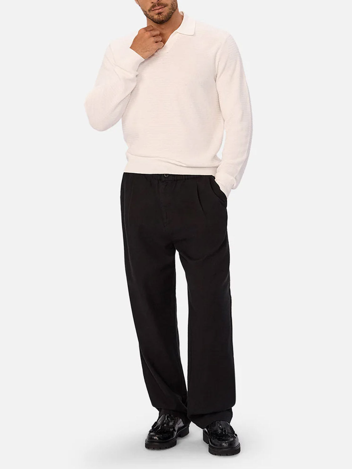 A man stands with one hand in his pocket, wearing a long-sleeve white polo shirt and Industrie’s The Relaxed Mallorca Pant in breathable cotton with a relaxed fit, paired with black dress shoes against a plain light gray background.