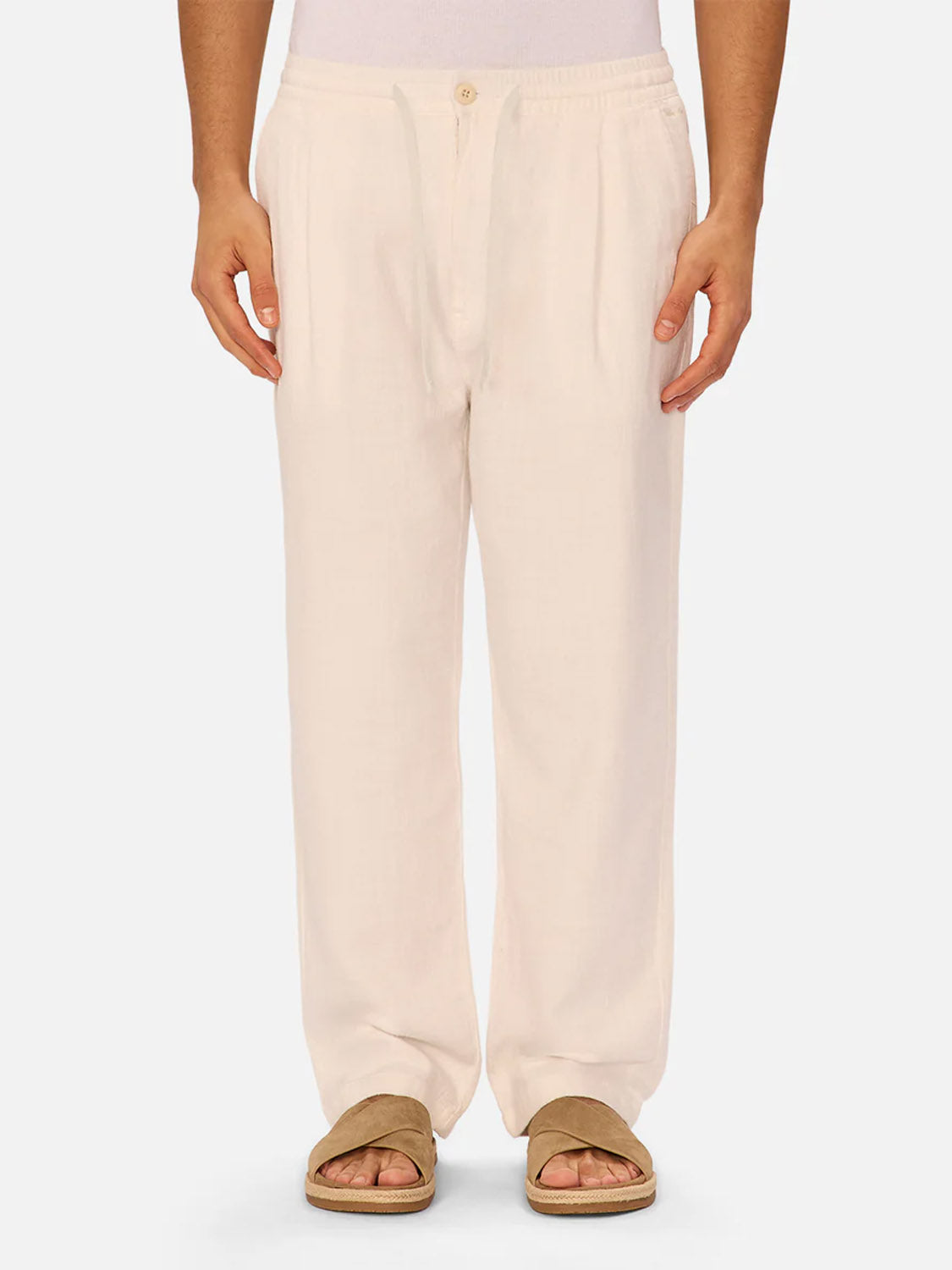 A person wearing The Relaxed Mallorca Pant by Industrie in light beige, crafted from breathable cotton with a relaxed fit, and brown slide sandals stands against a plain background, photographed from shoulders to feet.