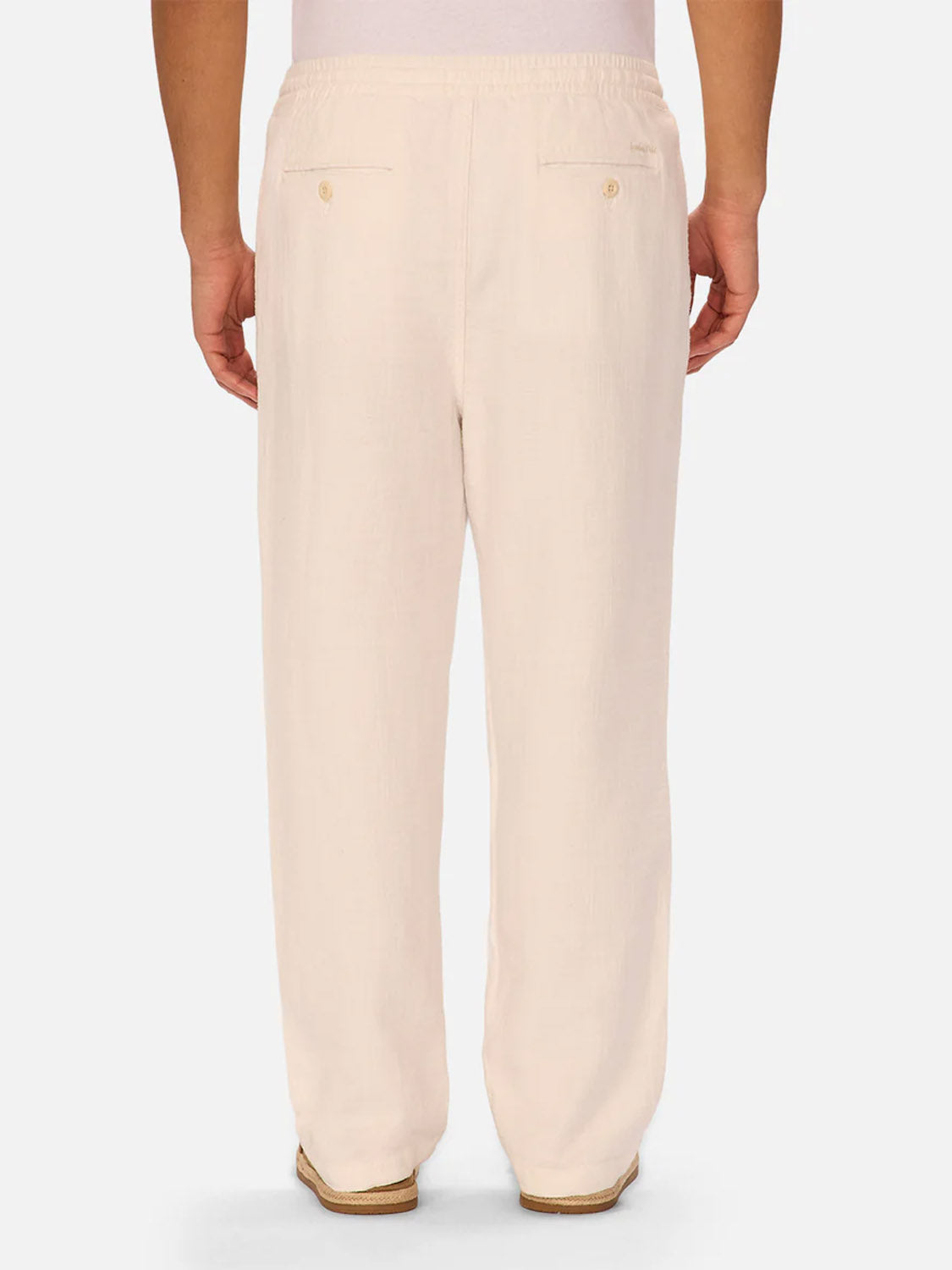 A person is seen from the back wearing Industrie’s The Relaxed Mallorca Pant in light beige. These cotton pants feature a relaxed fit, elastic waistband, two buttoned back pockets, and are set against a plain light background.