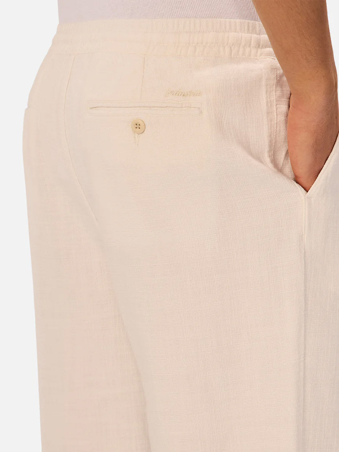 A close-up of a person wearing Industrie’s The Relaxed Mallorca Pant in light beige, crafted from breathable cotton with a relaxed fit, featuring a buttoned back pocket with embroidered logo and one hand in the side pocket.