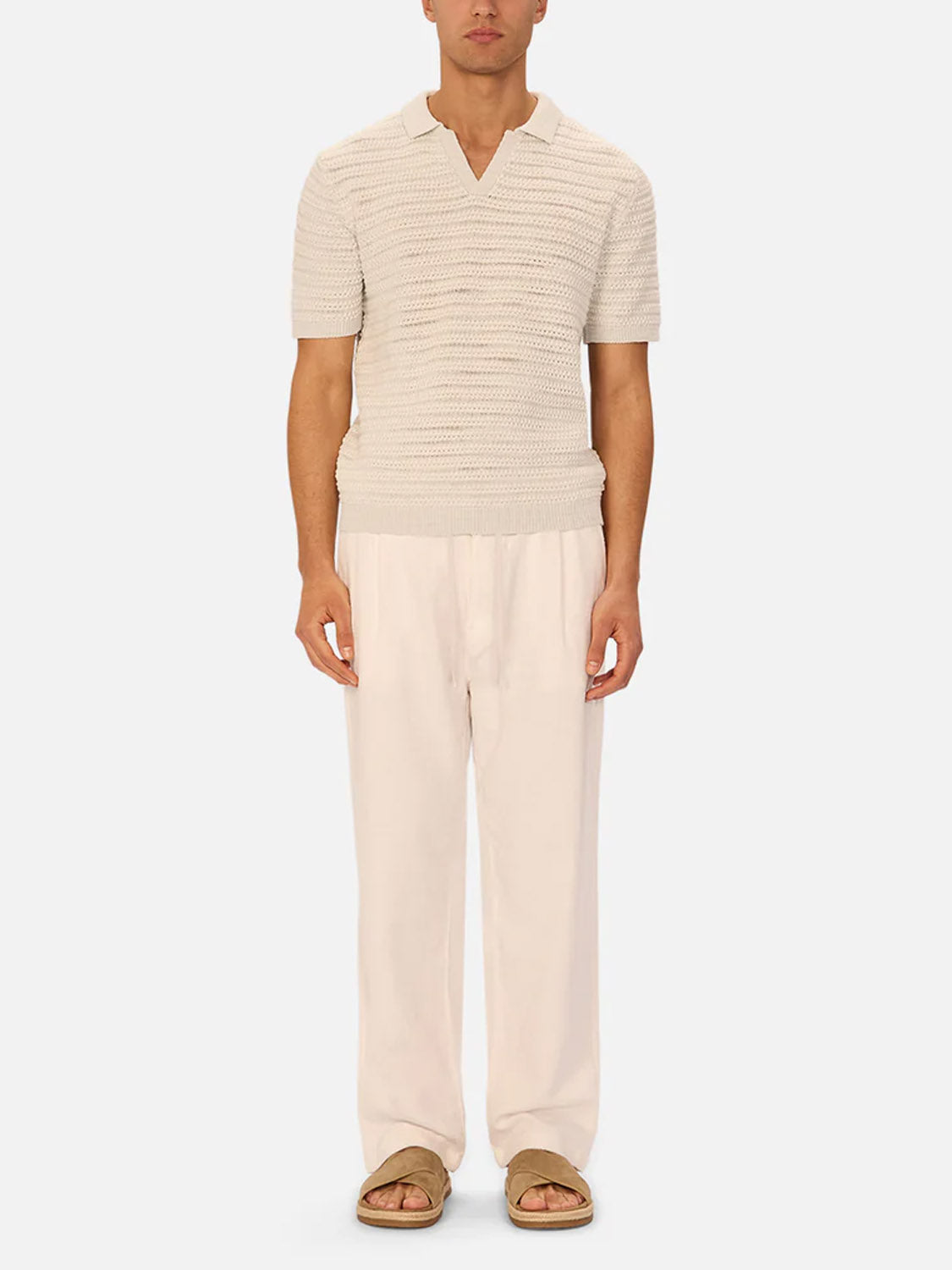 A man faces forward against a white background wearing a short-sleeve textured cream knit polo, Industrie’s The Relaxed Mallorca Pant in a relaxed fit, and tan slide sandals.
