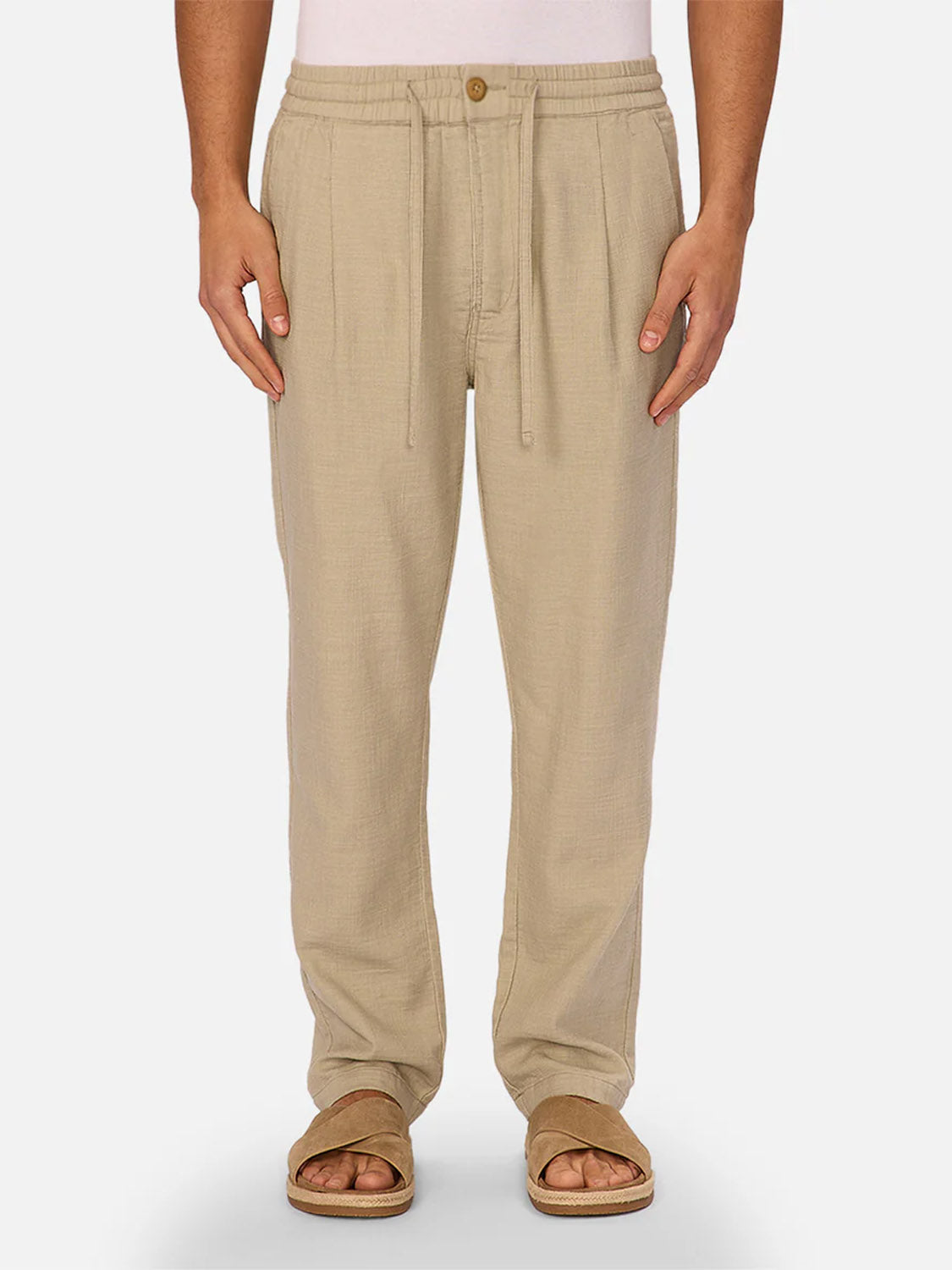 A person wears The Mallorca Pant by Industrie—light beige, breathable cotton pants with a drawstring and a brown button—paired with brown sandals. Only the lower half is visible against a plain light background.