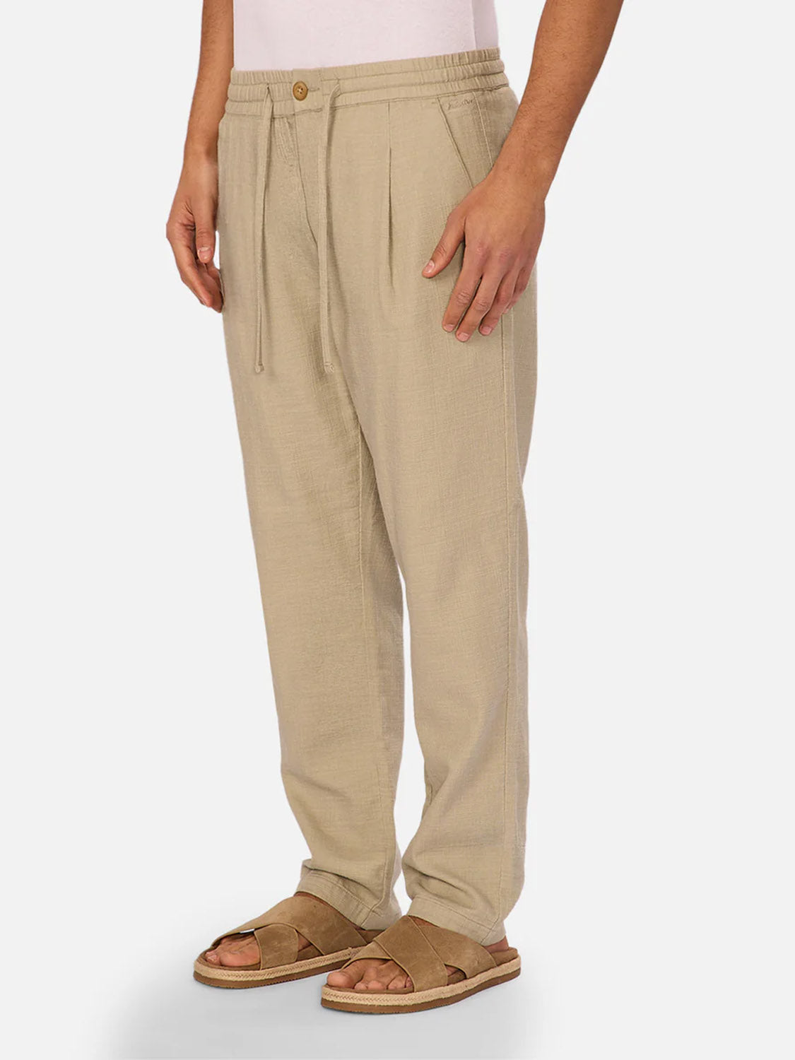 A person wears The Mallorca Pant by Industrie—loose-fitting, beige cotton pants with pleats—paired with brown sandals, standing against a plain white background.