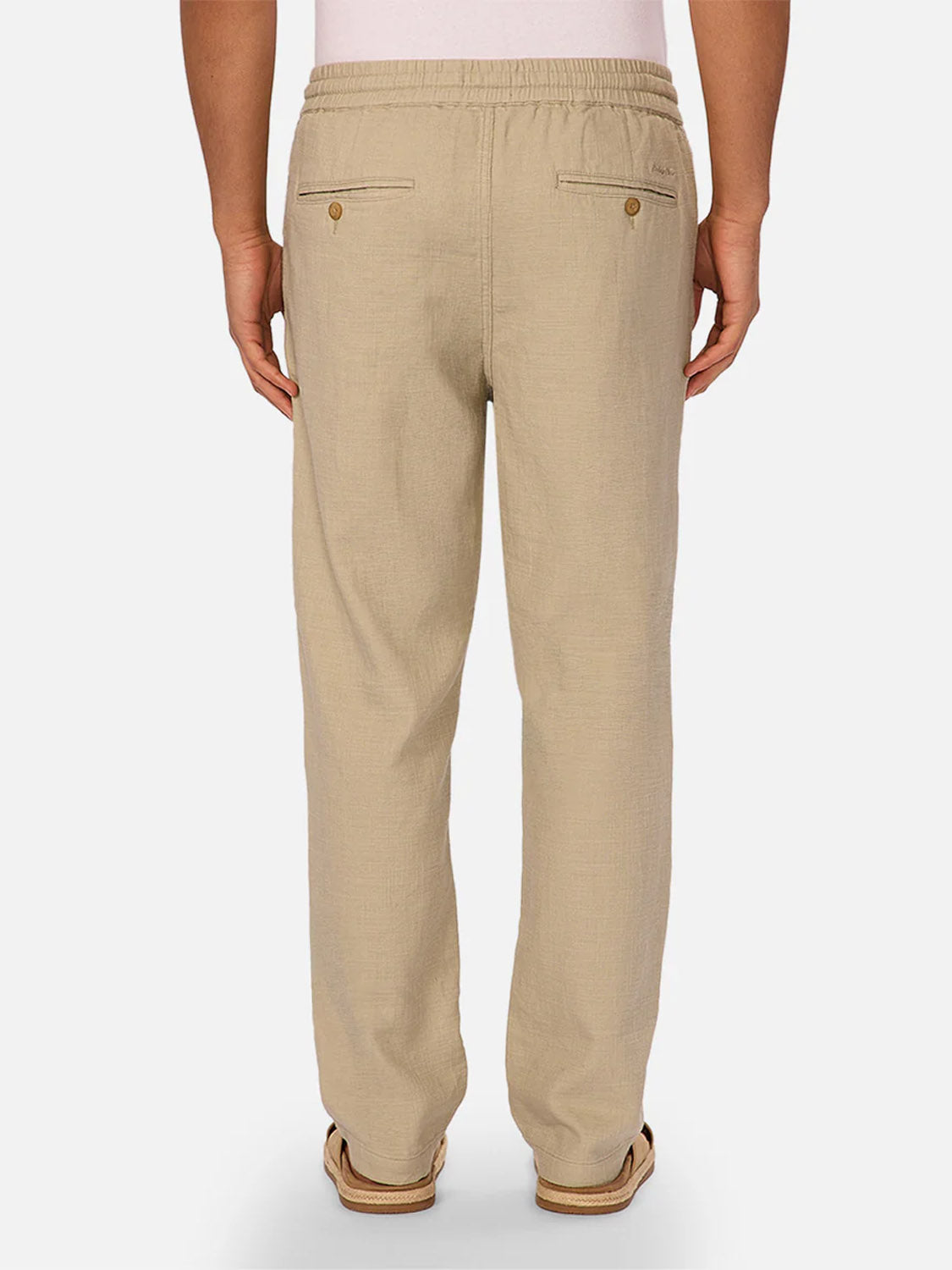 A person is shown from the back wearing The Mallorca Pant by Industrie in beige linen-cotton with an elastic waistband and two buttoned back pockets, paired with brown sandals. The background is plain white.
