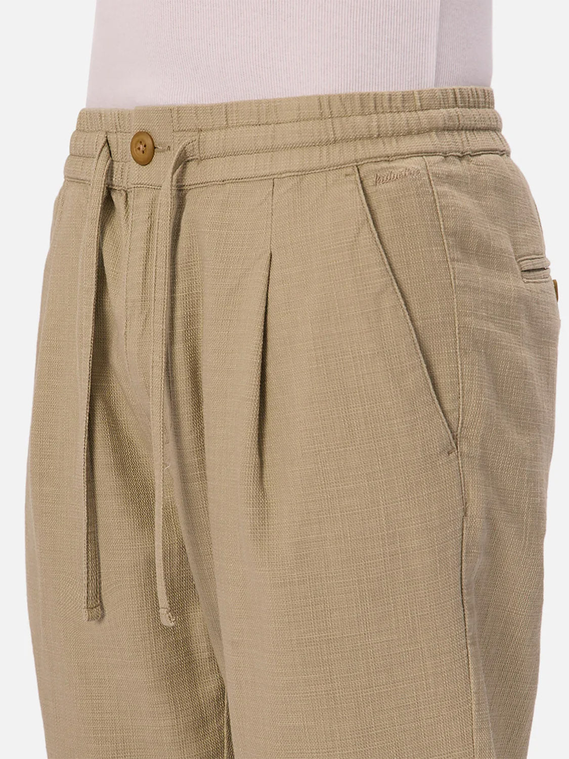 Close-up of a person wearing The Mallorca Pant by Industrie, featuring light beige, breathable cotton with a subtle textured pattern, elastic waistband, button closure, and side pockets. Styled with a white top.