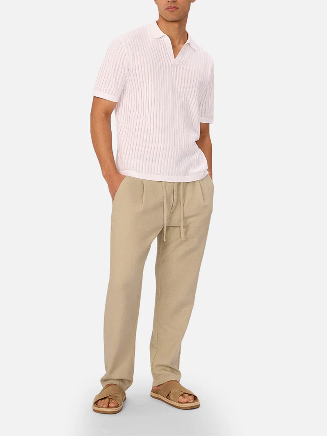 A man stands with hands in pockets, wearing The Mallorca Pant by Industrie—breathable beige drawstring linen pants—paired with a pale pink short-sleeve knit polo shirt and brown sandals against a plain light gray background.
