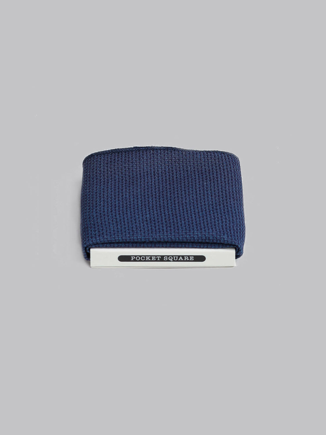 A neatly folded navy blue Malmo Pocket Square by ID Menswear, handmade in Brooklyn and secured in a holder marked POCKET SQUARE, sits against a plain light gray background.