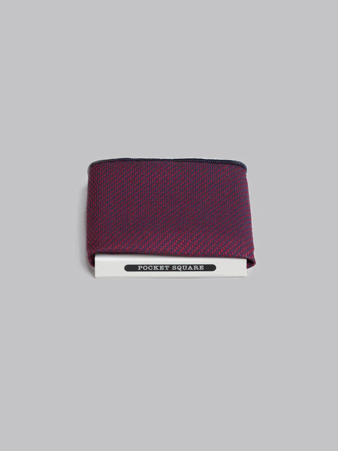 A folded Malmo Pocket Square by ID Menswear, handmade in Brooklyn from 100% cotton with a red and navy pattern, rests in white packaging labeled POCKET SQUARE on a plain light gray background.
