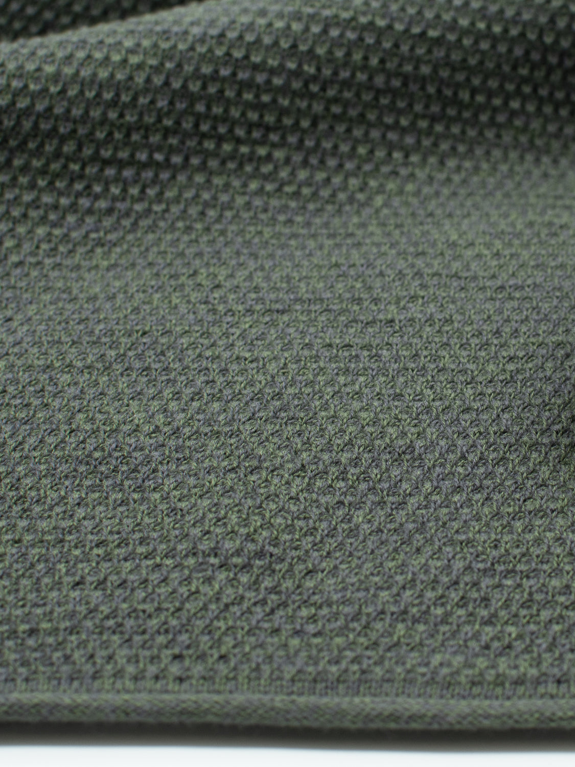 A close-up of ID Menswears Malta Rolled Neck Sweater Tee reveals a textured, muted earthy-green fabric with a small geometric pattern. Its slightly coarse appearance and subtle shading add depth and dimension.