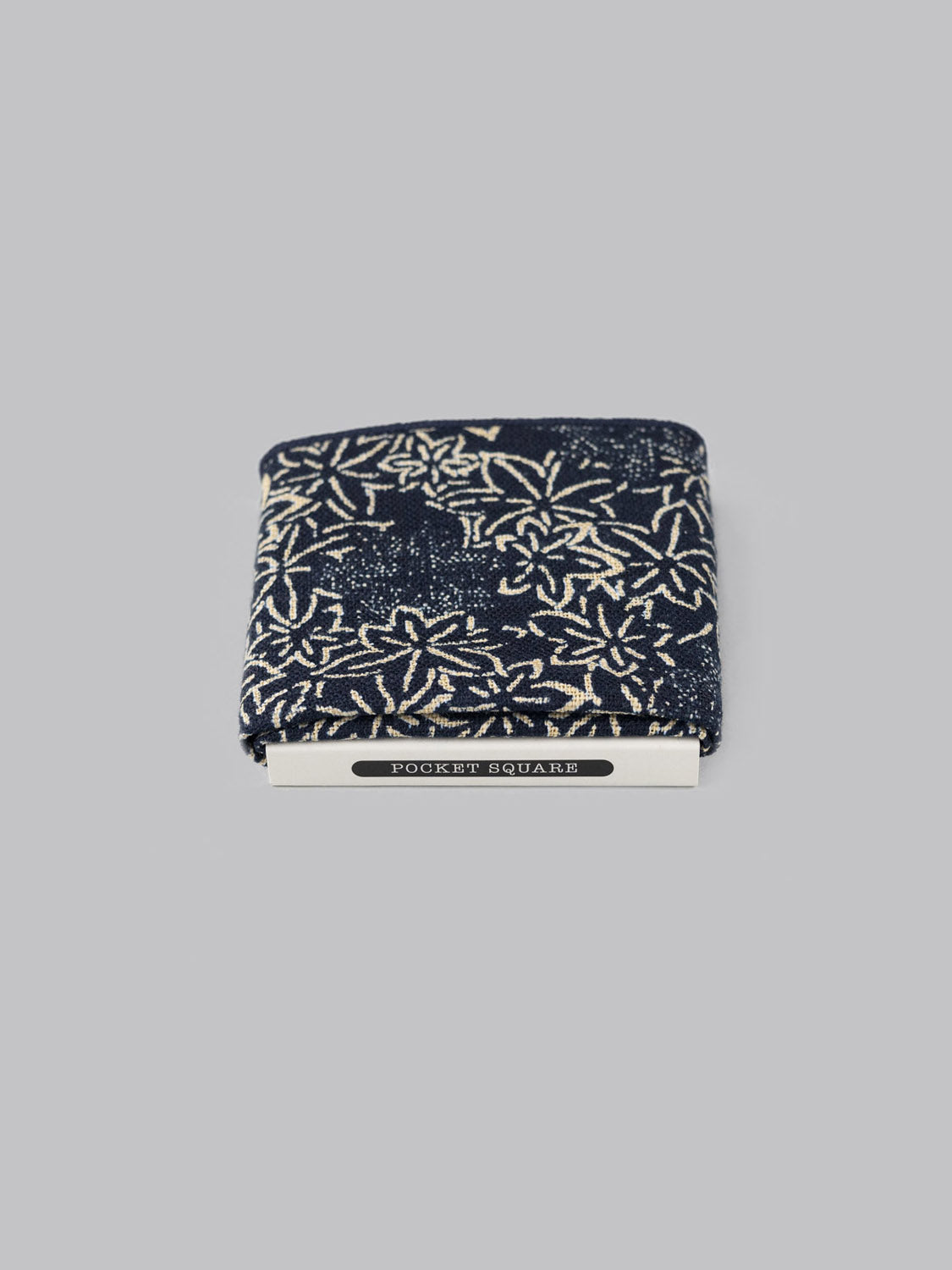 The Maple Pocket Square by ID Menswear is a dark blue cotton accessory with a white floral pattern, handmade in Brooklyn. Its neatly folded in a white holder labeled POCKET SQUARES against a plain gray background.