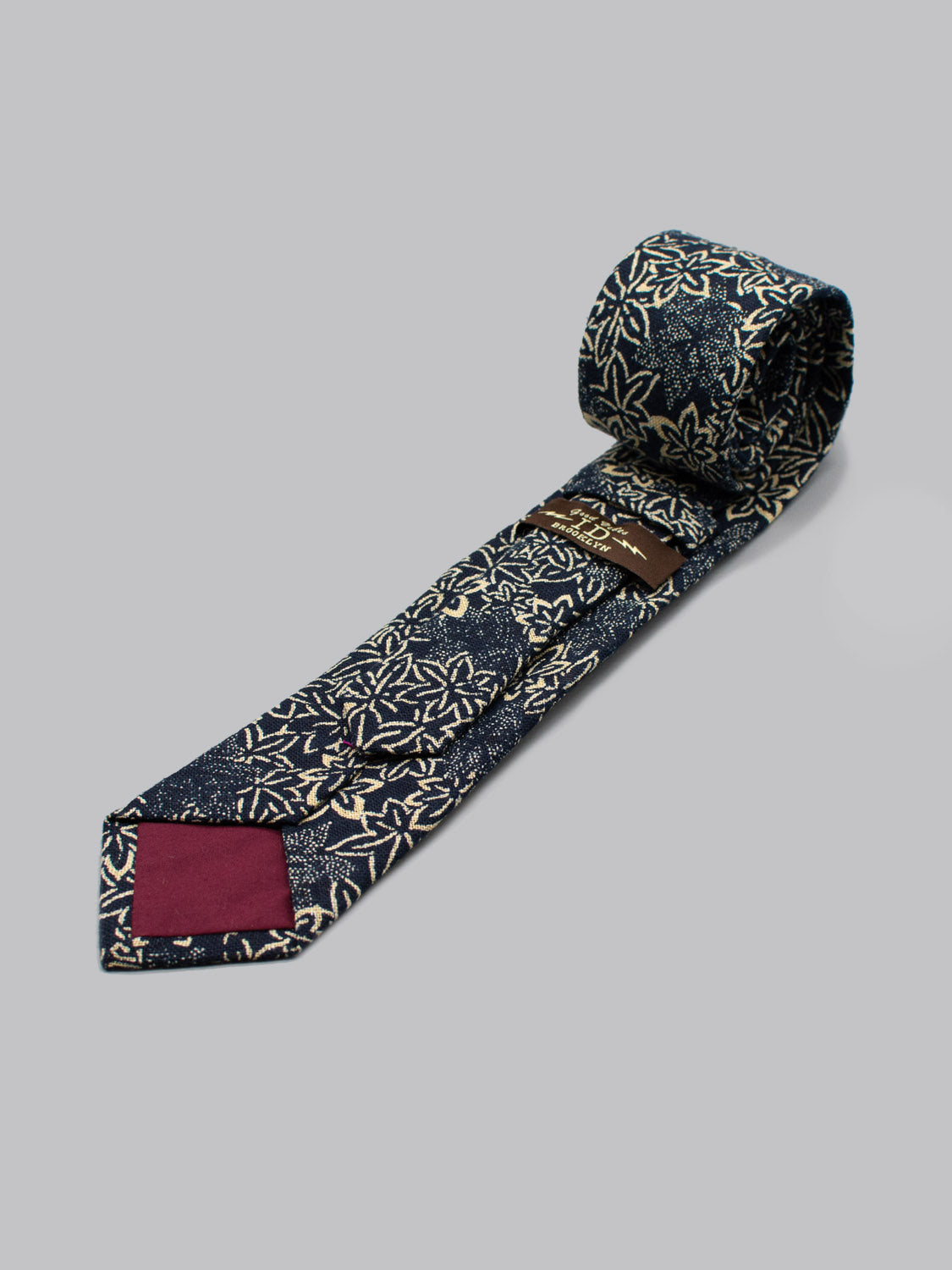 The Japanese Printed Maple Tie by ID Menswear, a rolled-up navy blue necktie with an off-white floral pattern and maroon tip lining, displays classic Brooklyn style on a plain light grey background.