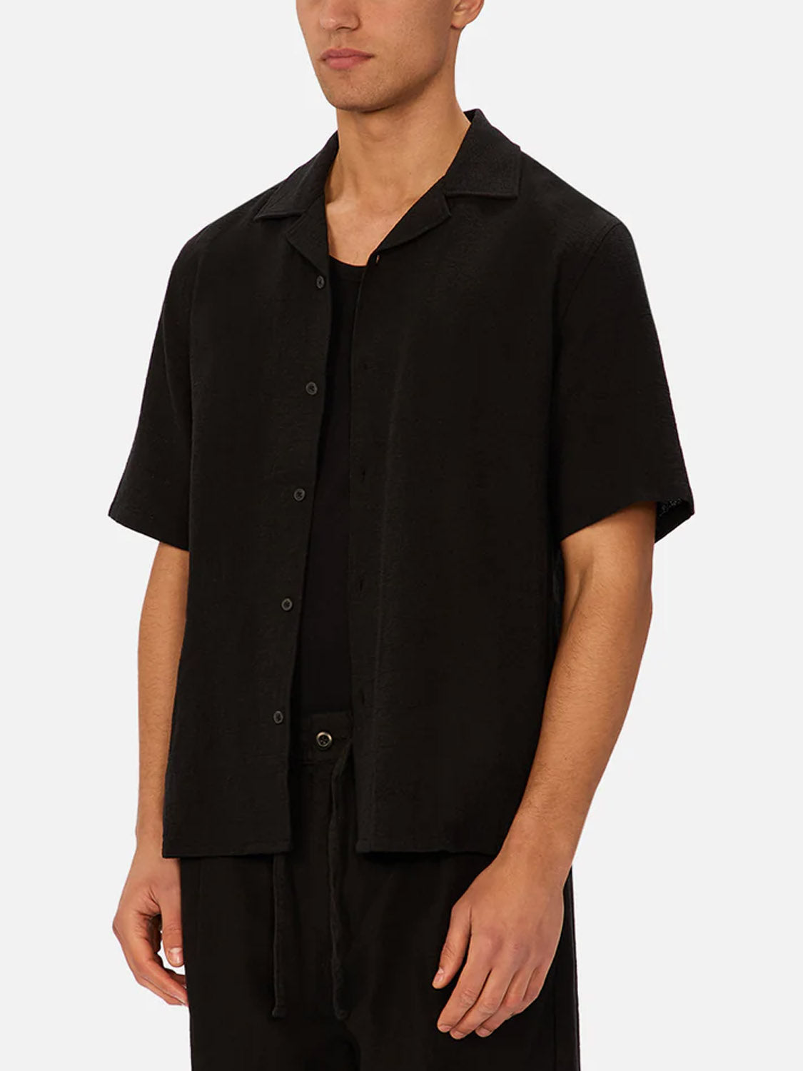 A person, face partially out of frame, stands against a plain light background wearing The Marbella Short Sleeve Shirt by Industrie in black over a matching top and pants.