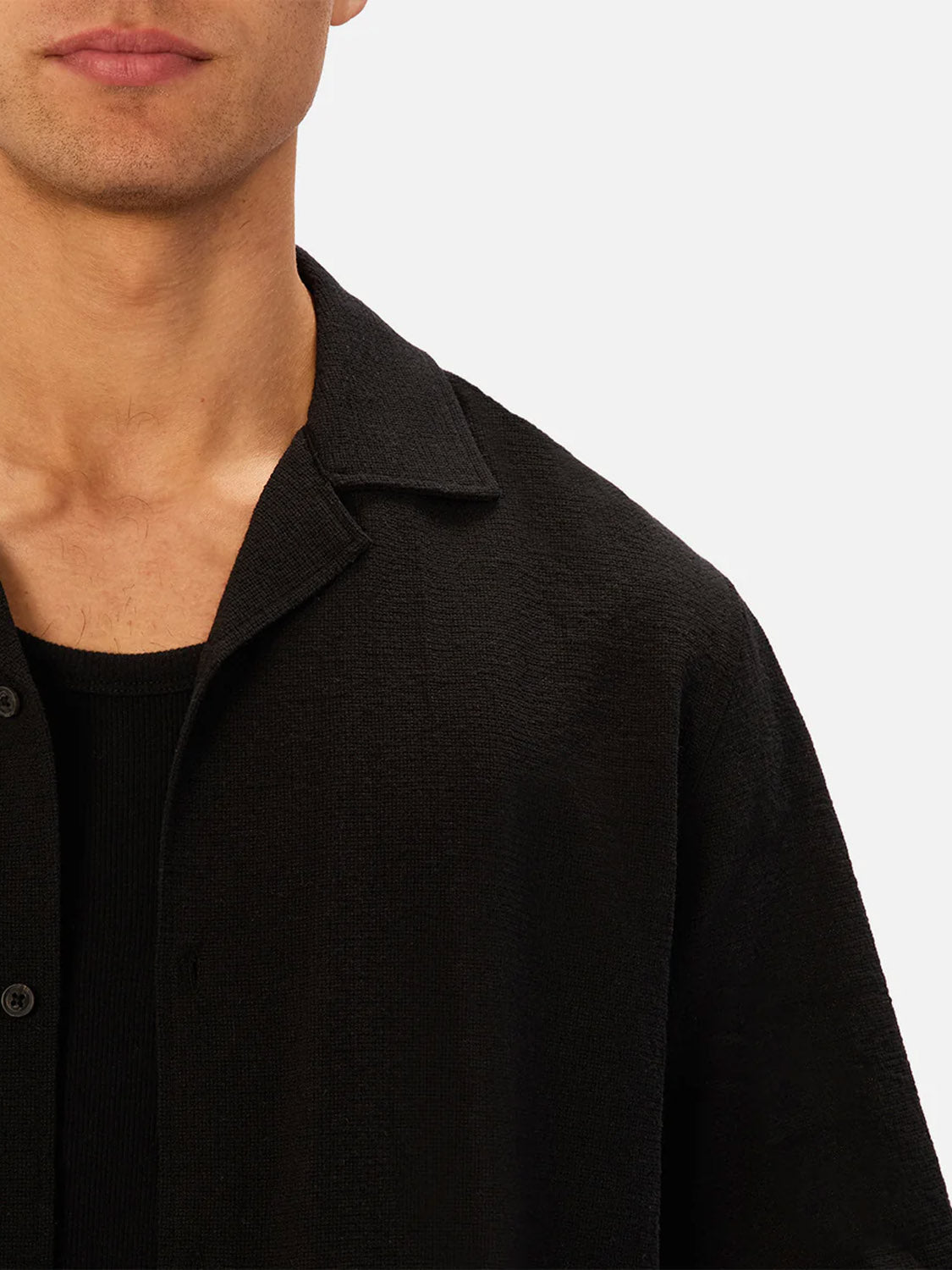 A close-up shows the lower face and upper torso of a person wearing Industrie’s The Marbella Short Sleeve Shirt in black, layered over a black top, set against a plain light background.