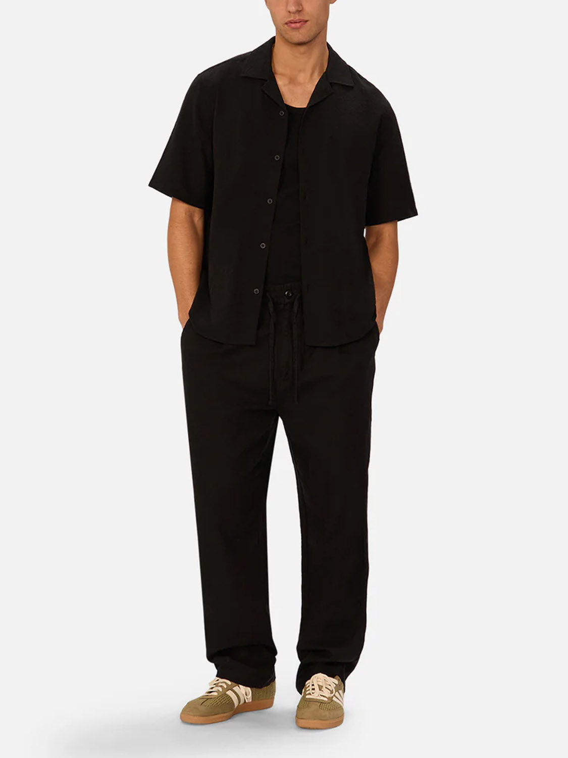 A man stands with his hands in his pockets against a plain background, wearing Industrie’s Marbella Short Sleeve Shirt—lightweight and black—over a black tee, loose drawstring black pants, and tan sneakers with white accents.