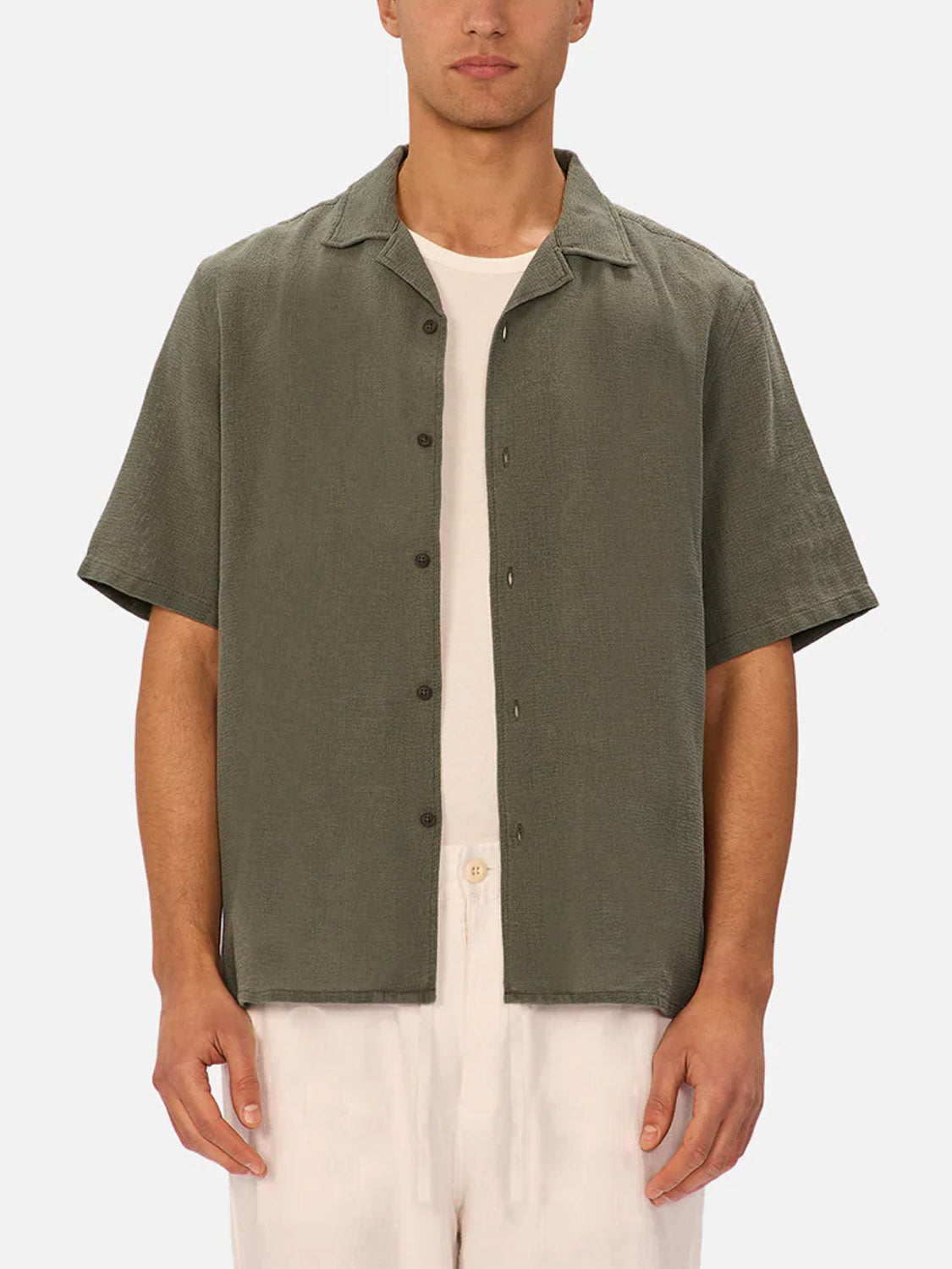 A man stands against a plain light background wearing the Industrie Marbella Short Sleeve Shirt in olive green over a white t-shirt, paired with light-colored pants.