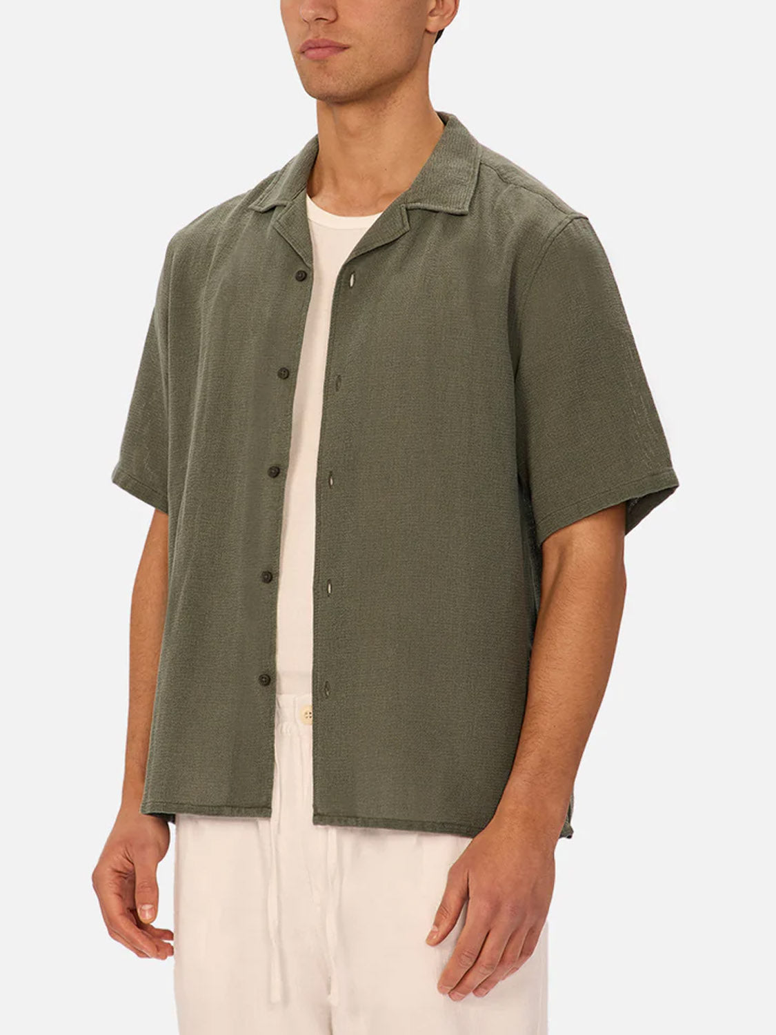 A man wears The Marbella Short Sleeve Shirt by Industrie in olive green over a white T-shirt and white drawstring pants, standing against a plain light background.