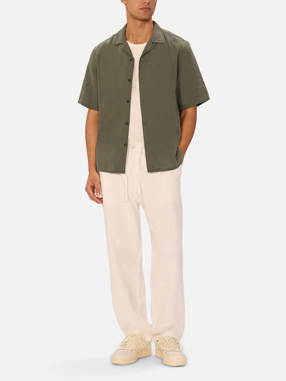A man wears the Industrie Marbella Short Sleeve Shirt in olive green over a white tee, paired with cream drawstring pants and sneakers, with hands in pockets against a plain light background.