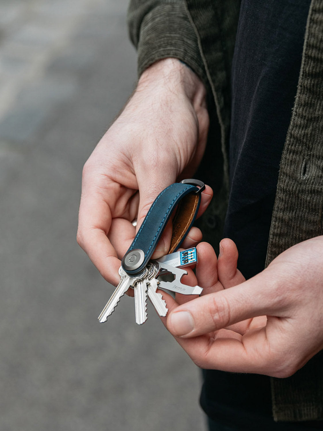 A person holds an Orbitkeys Key Organizer Crazy-Horse Cowhide Leather in blue and black, with several keys and a small multitool attached. The background is a blurred street surface.