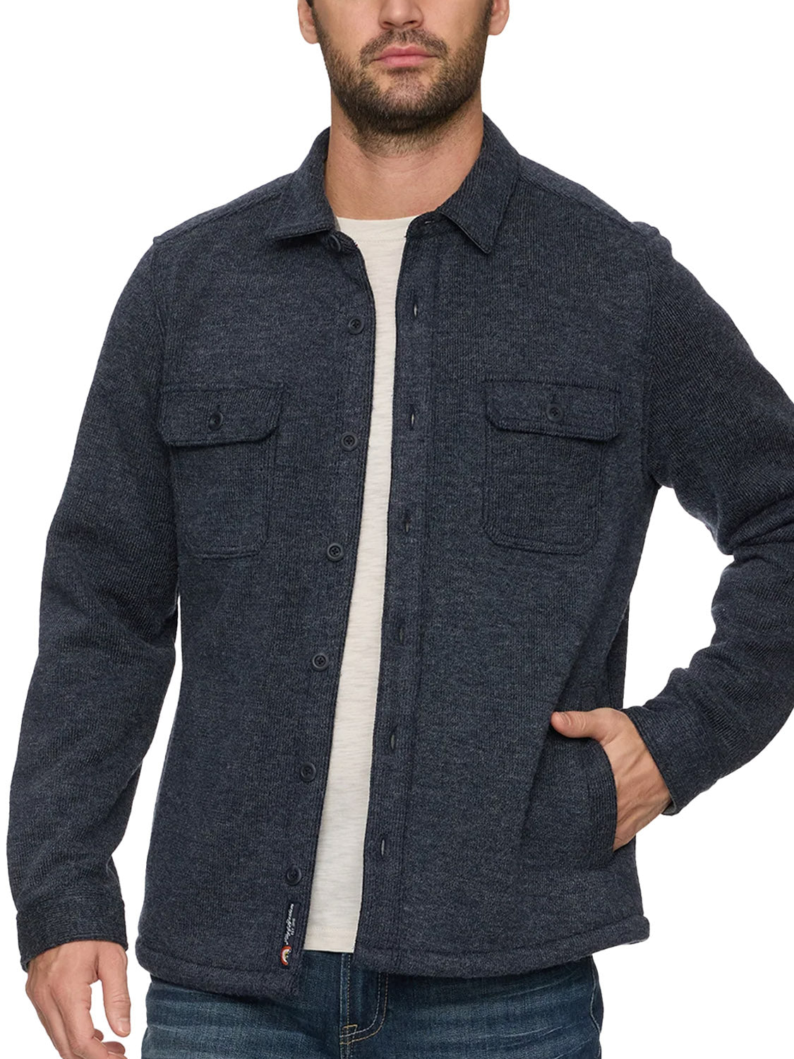 A man wears the Flag and Anthem Mason Fleece Lined Shirt Jacket, featuring a dark blue hue, two chest pockets, and a cozy fleece interior, layered over a light shirt and blue jeans with one hand in his pocket.
