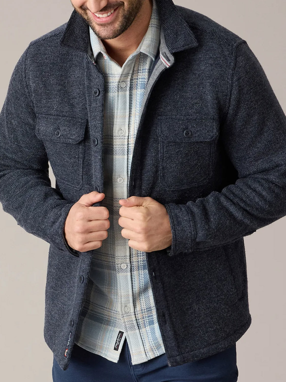 A man smiles as he buttons the Flag and Anthem Mason Fleece Lined Shirt Jacket over a plaid shirt, shown from shoulders to hips with both hands adjusting the jacket—an ideal men’s outerwear choice against a neutral background.
