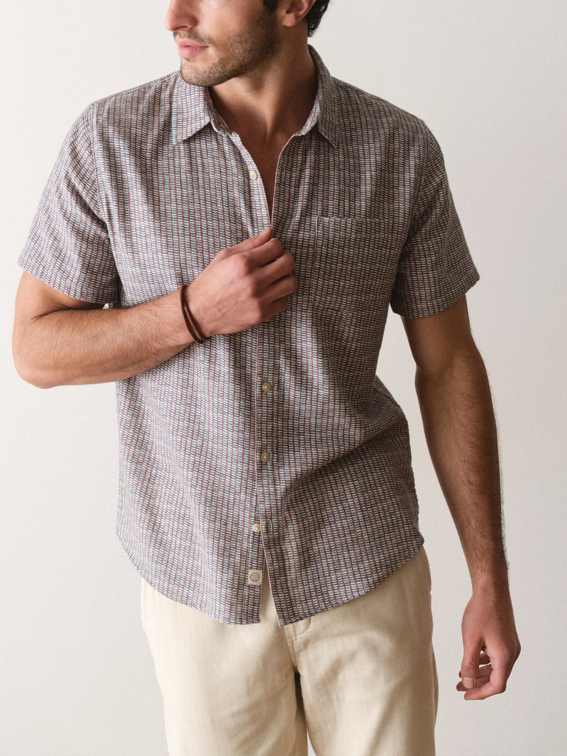 A man in a Marine Layer Stretch Selvage Short Sleeve Shirt and light pants stands against a plain background, looking to the side while adjusting his shirt.